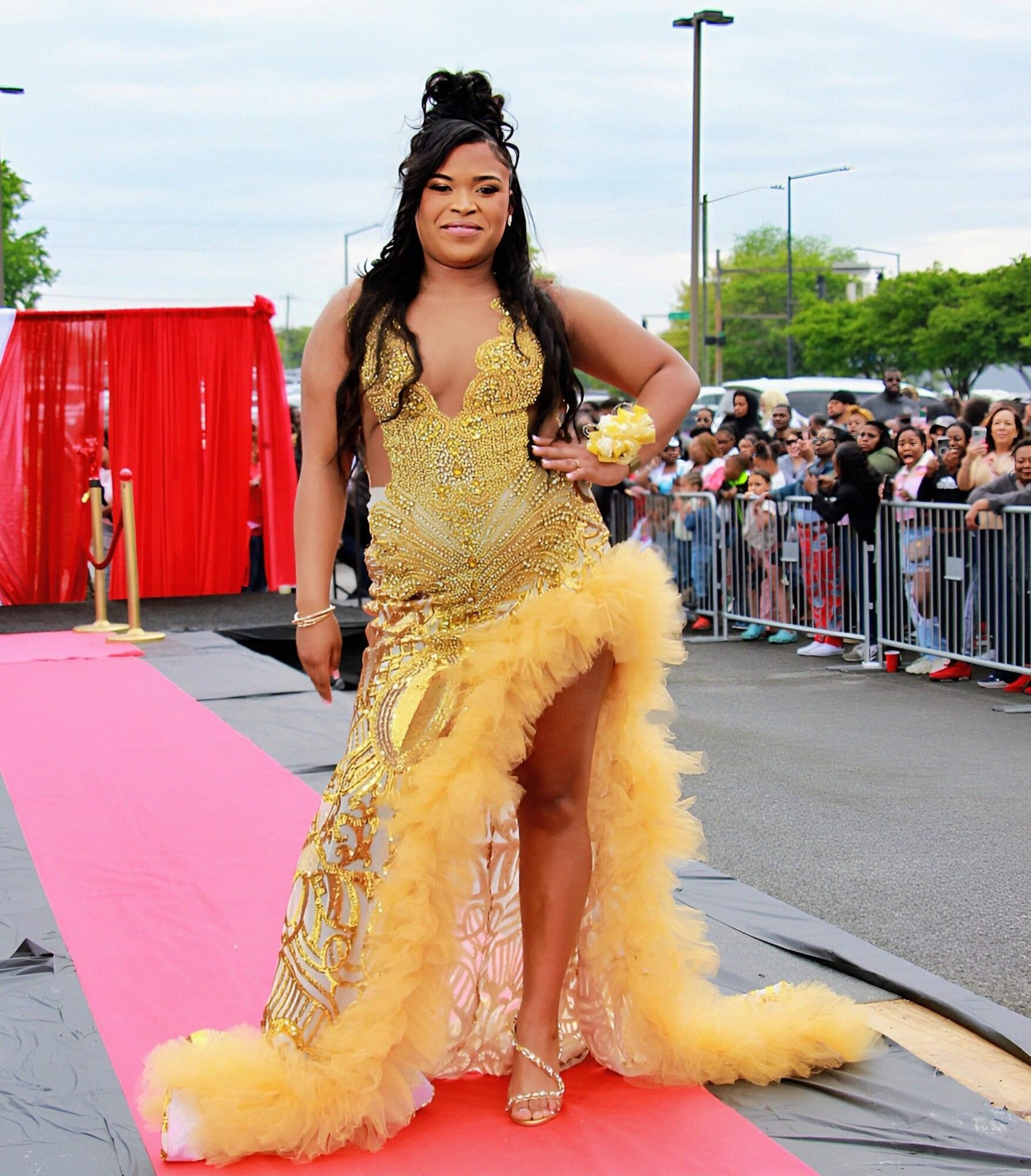 Woman in a yellow gown with a high slit, standing on a pink carpet outdoors, with a crowd behind barriers.