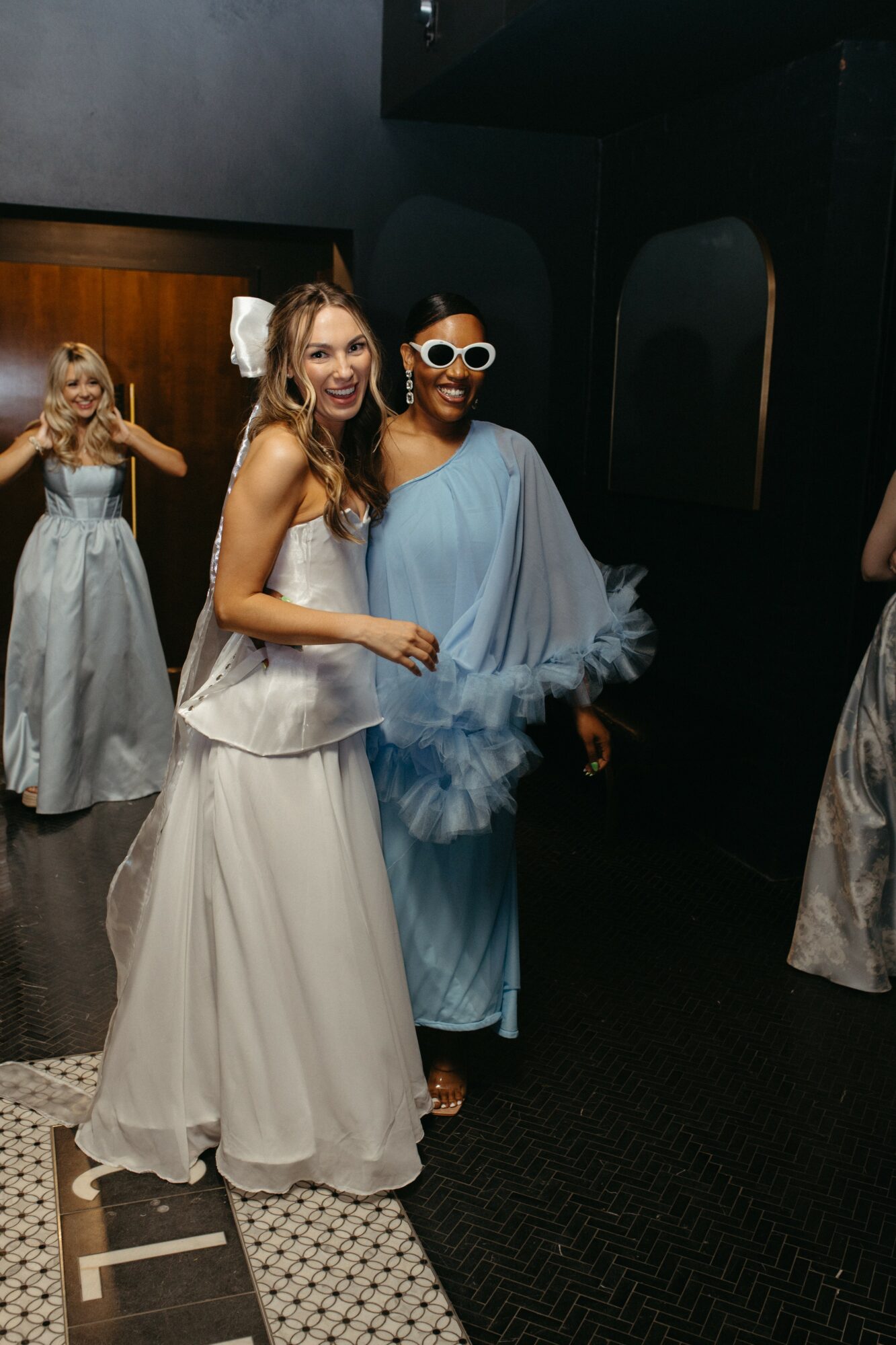 Two women in elegant dresses pose together, one in a white gown and the other in a blue dress with sunglasses, in a dark room.