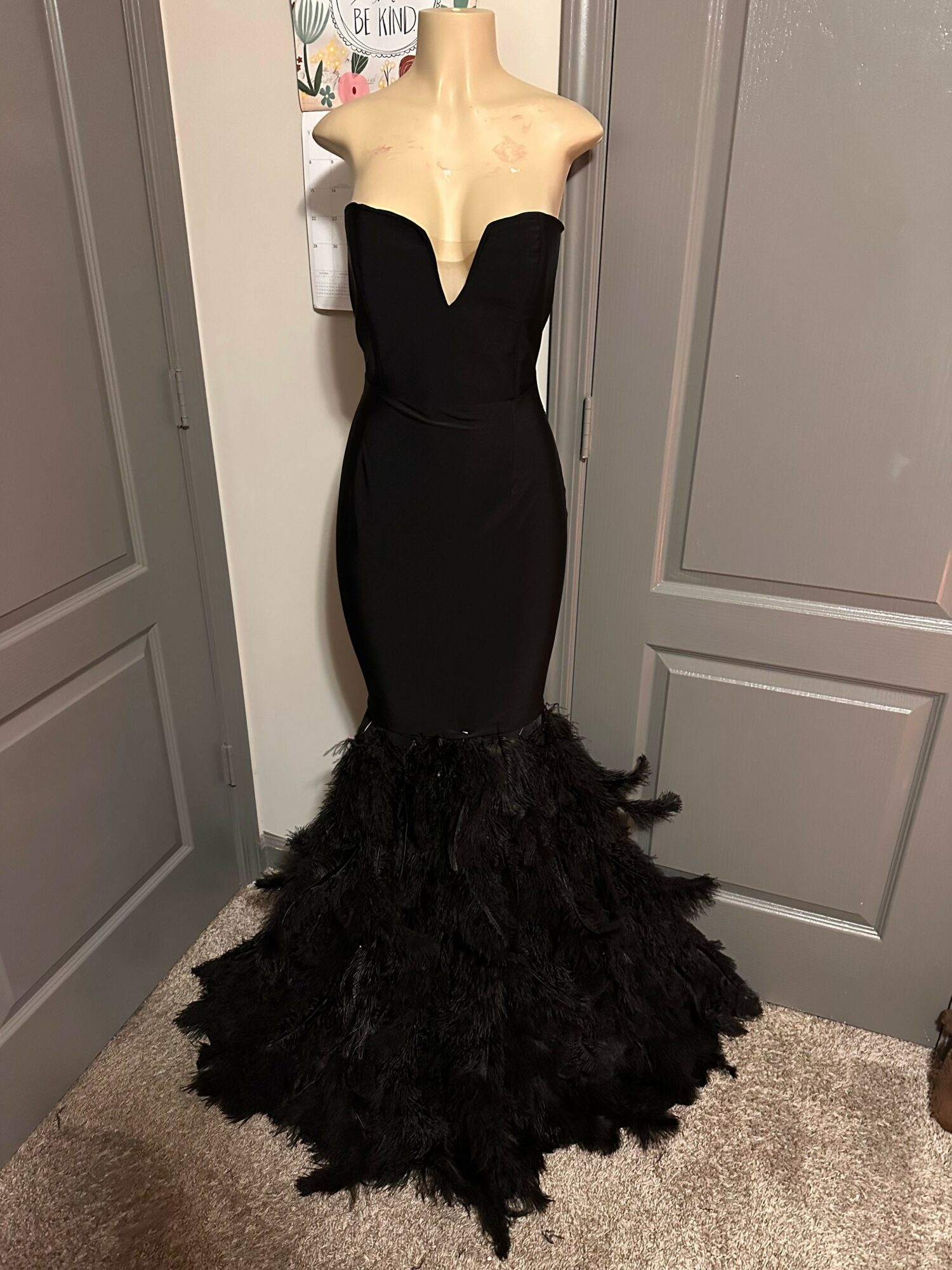 Black dress on mannequin with long, textured black hair, standing in front of gray doors, on carpeted floor.