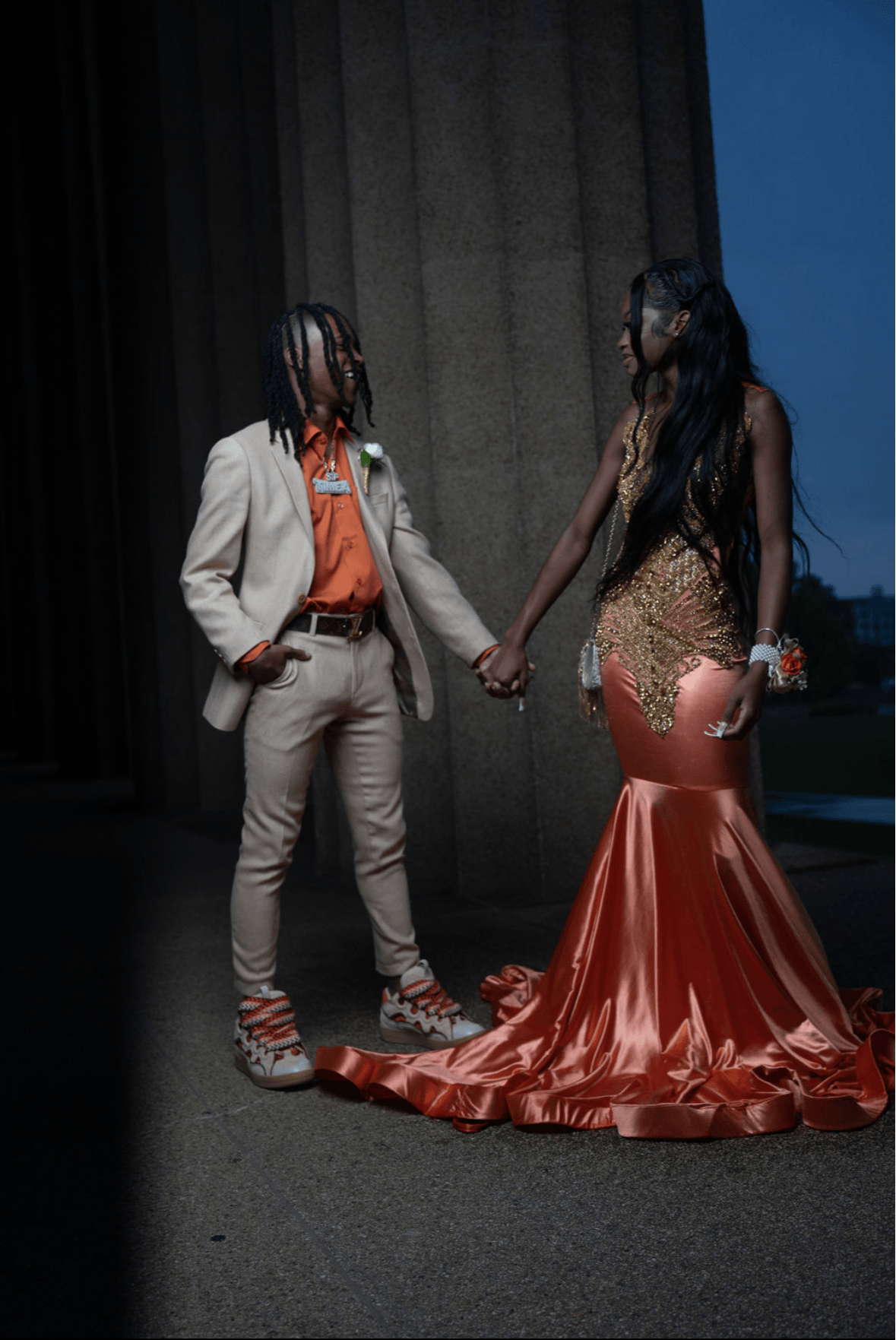 Two women holding hands, one in a beige suit and the other in a gold and orange gown, standing on a stage.