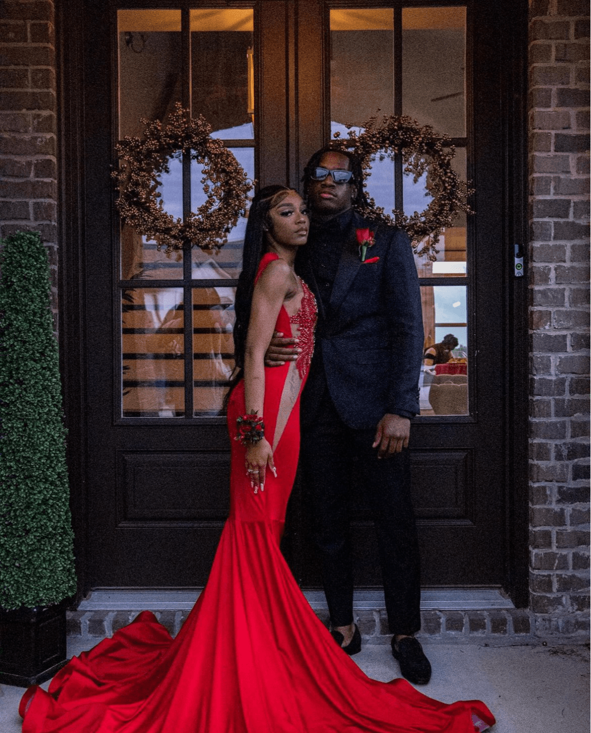 A couple stands in front of a door with two wreaths, woman in a red gown and man in a dark suit, nighttime setting.