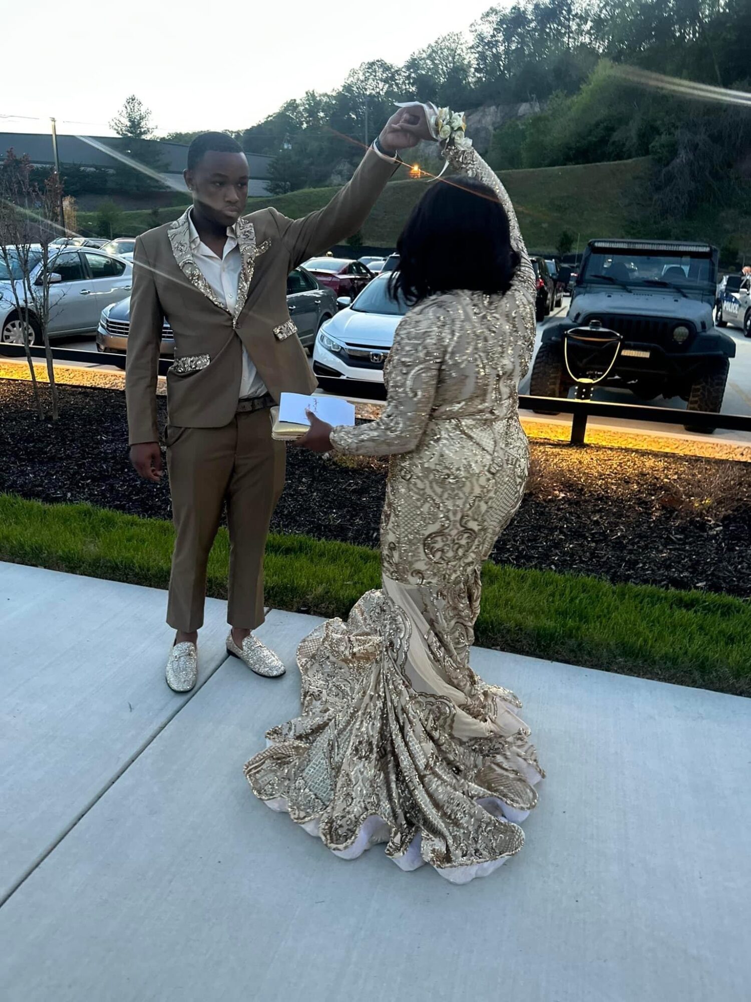 Two women in formal attire exchange a small object outdoors, with parked cars and greenery in the background.