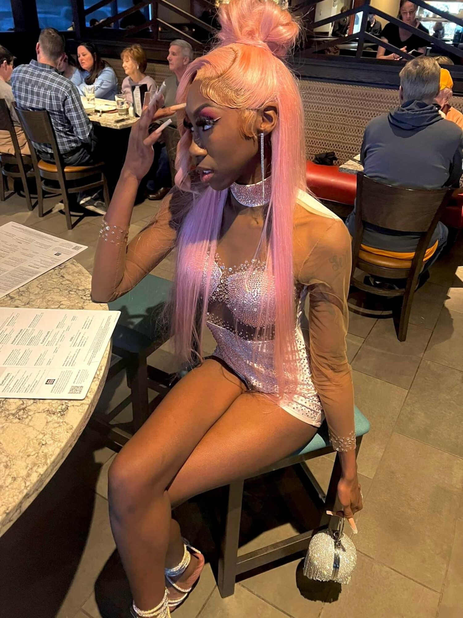 Person with long pink hair, sitting at a table in a restaurant, wearing a shiny top and shorts, holding a small bag.