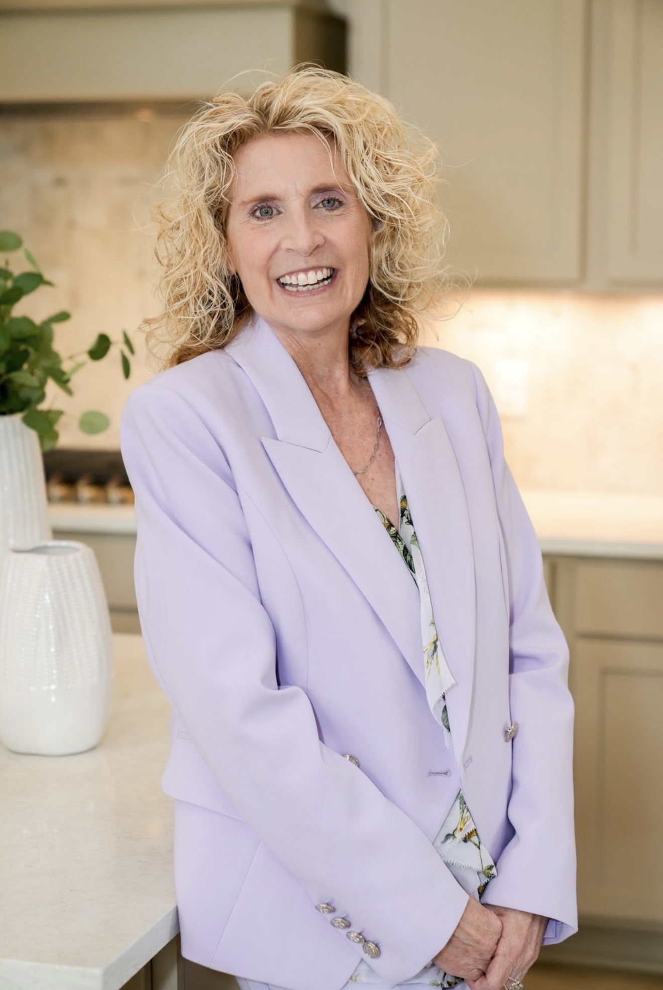 Smiling woman with curly blonde hair in a light purple blazer in a kitchen.