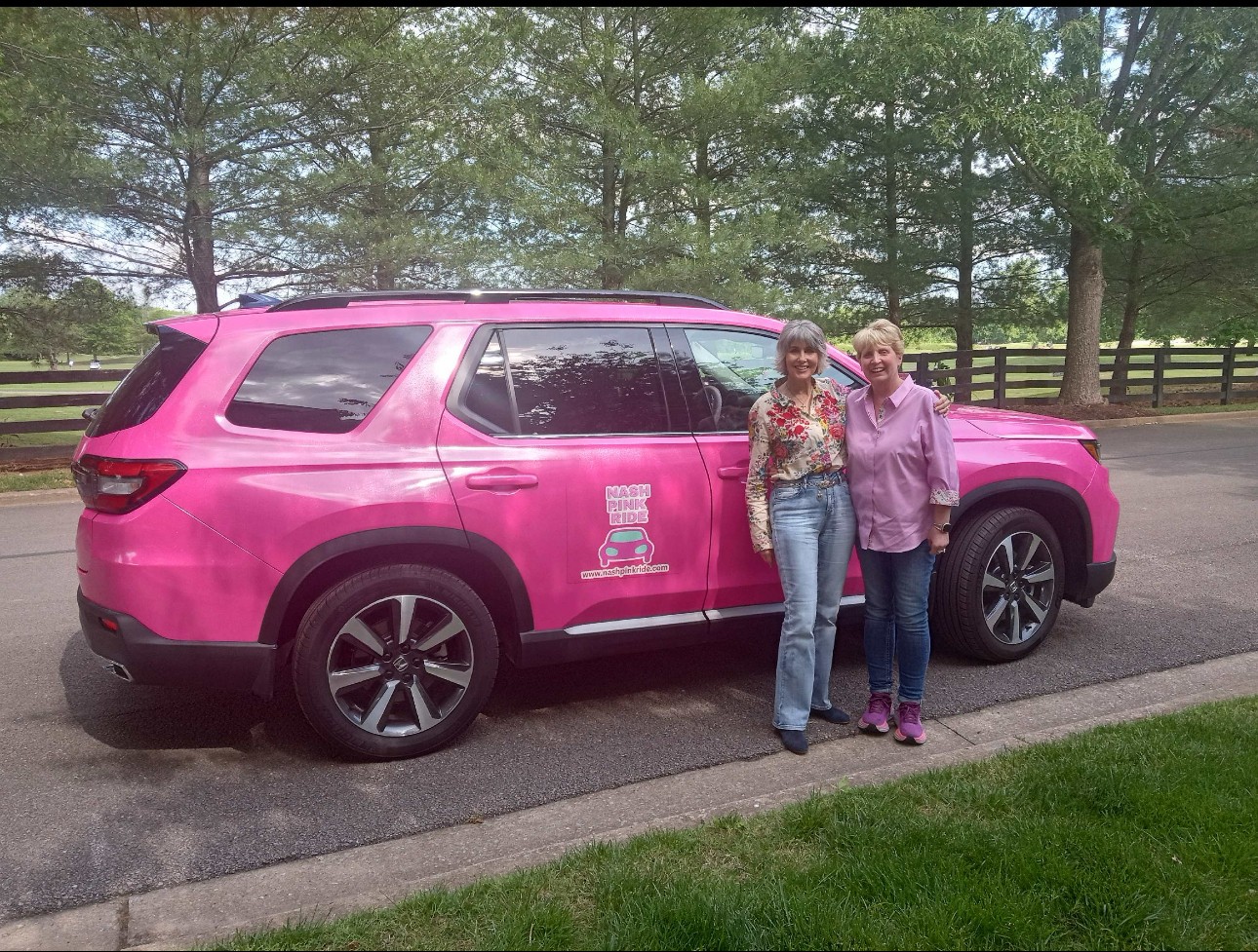 Two women stand next to a pink SUV on a paved road with trees and a fence in the background.