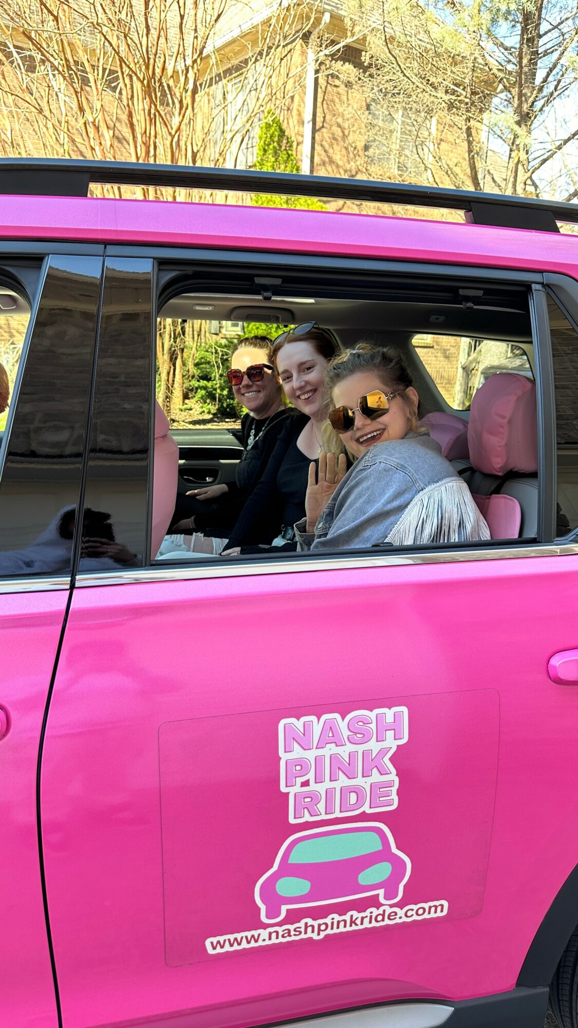 Three people sitting in a pink vehicle with a sticker that reads 'Nash Pink Ride' and a website URL, outdoors.