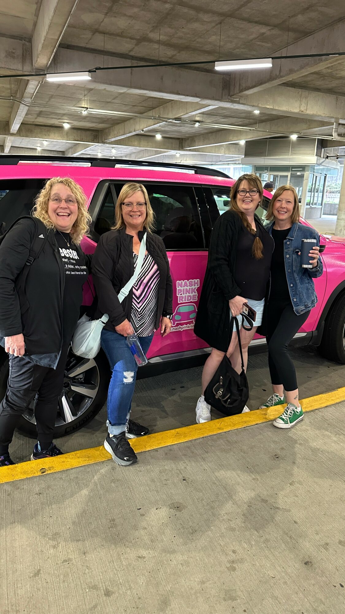 Four women standing in a parking garage next to a pink and black vehicle, smiling and holding bags.
