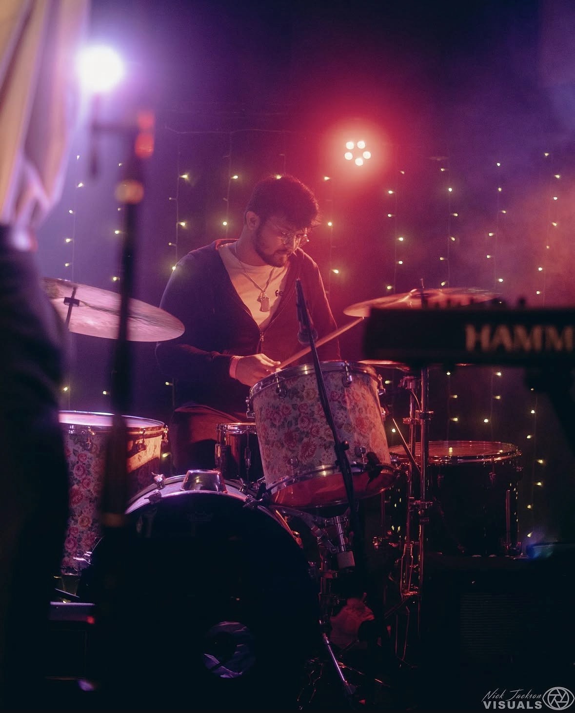 A person playing drums on stage with colorful lights and a keyboard nearby, in a concert setting.