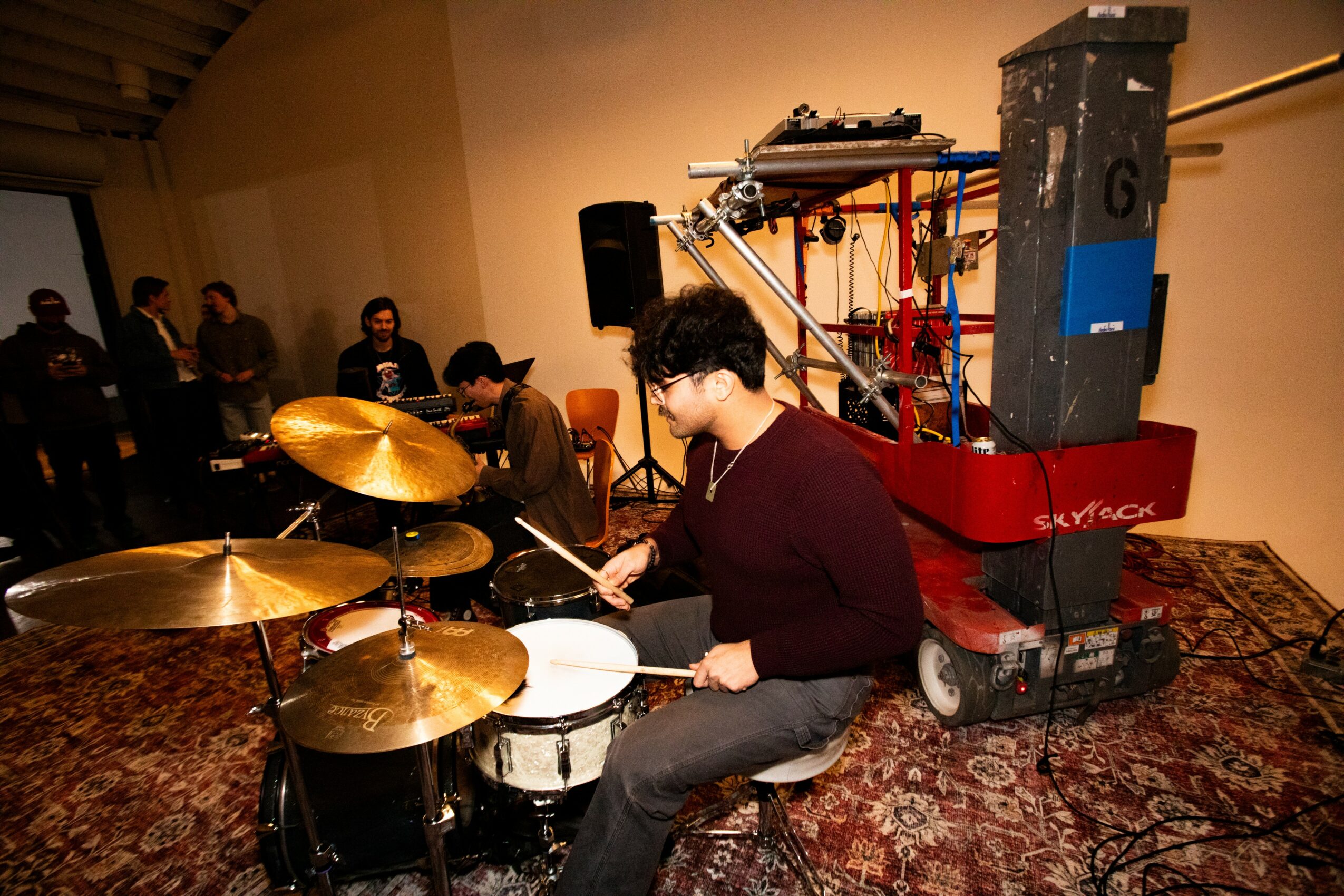Person playing drums in a room with audience and musical equipment, including a large speaker and colorful cables.