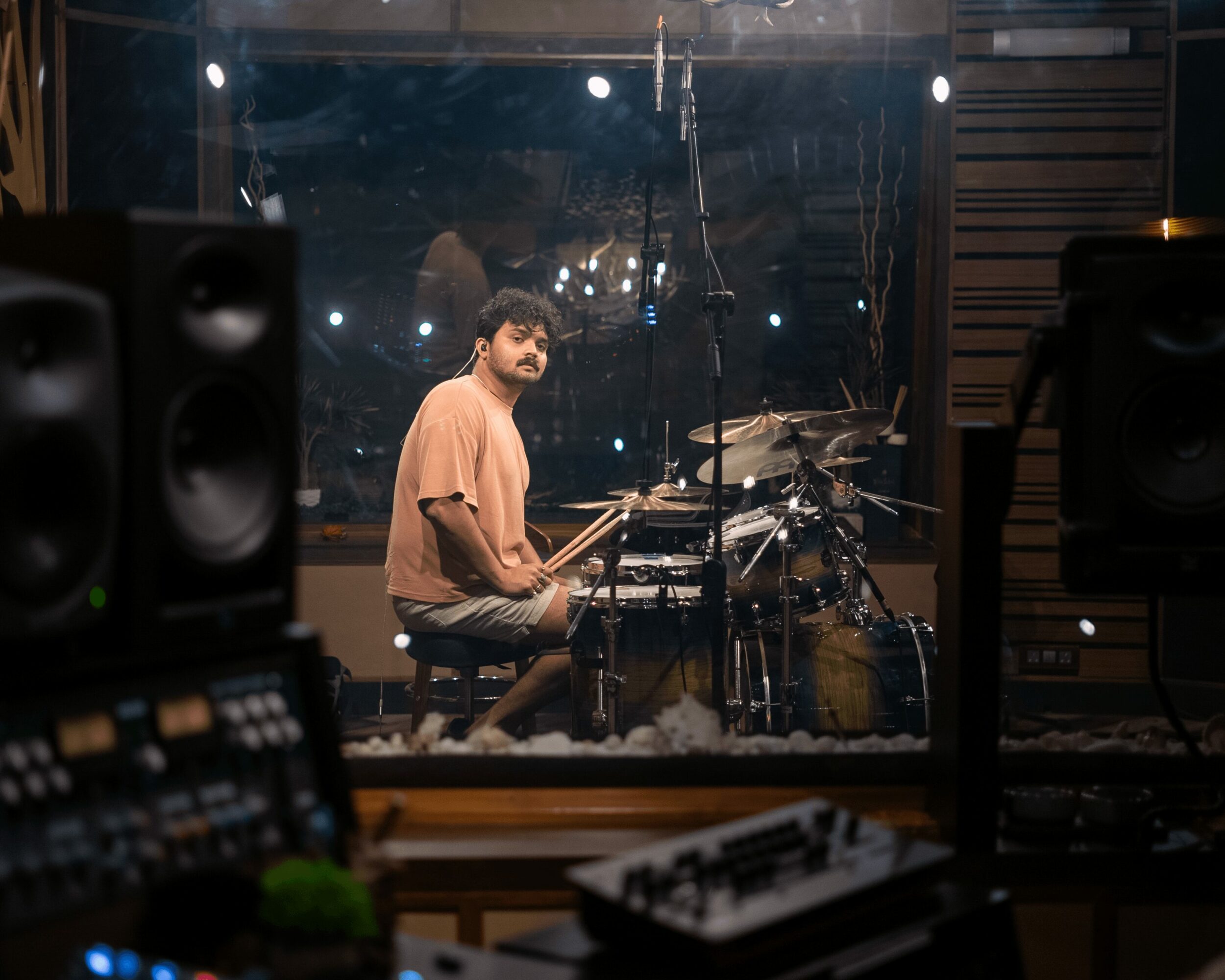 Musician playing drums in a recording studio, viewed through a glass window with audio equipment in foreground.