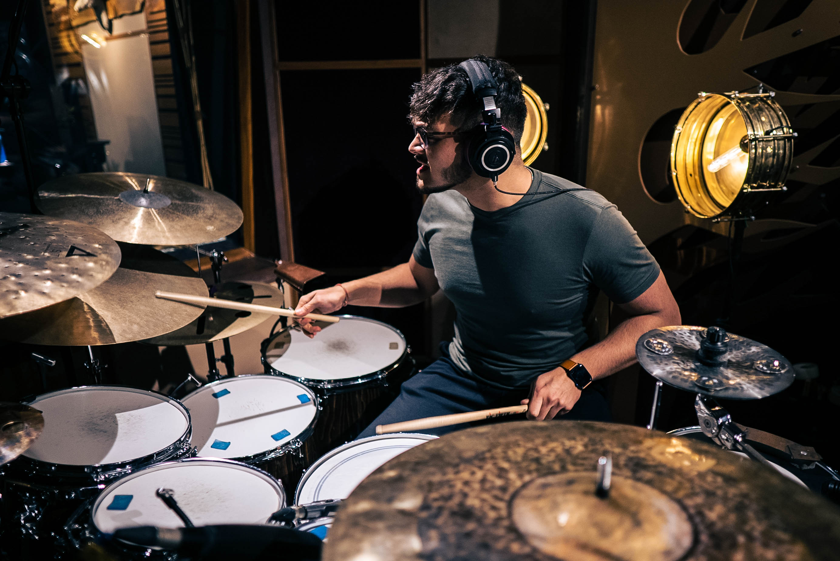 Person playing drums in a recording studio, wearing headphones and glasses, surrounded by drum equipment and studio lights.