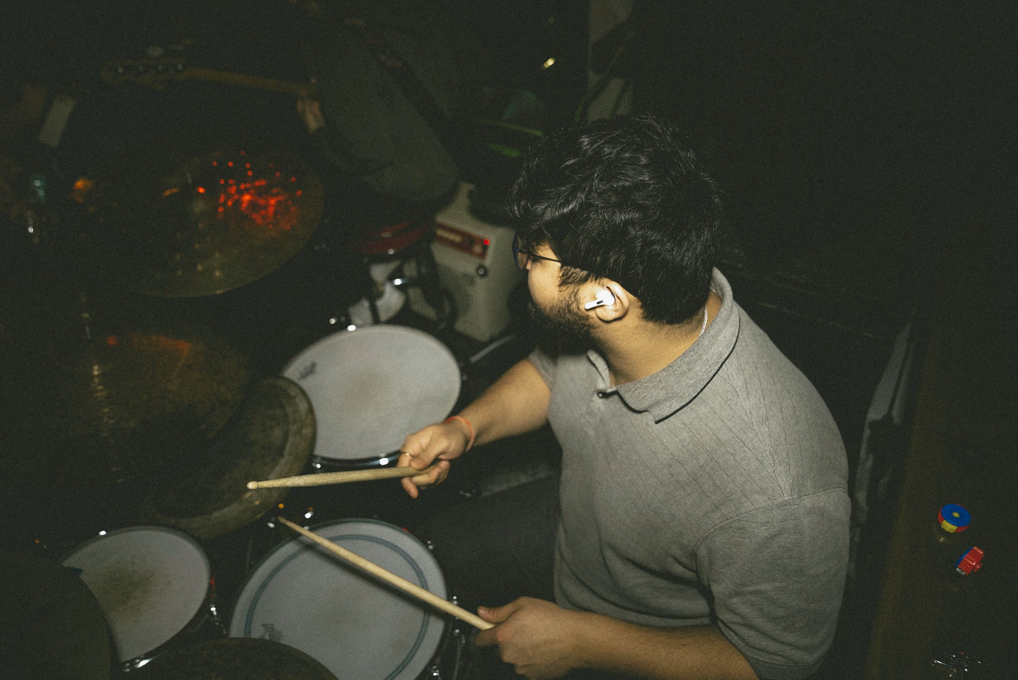 Person playing drums with drumsticks in a dark setting, wearing a gray shirt, viewed from the side.