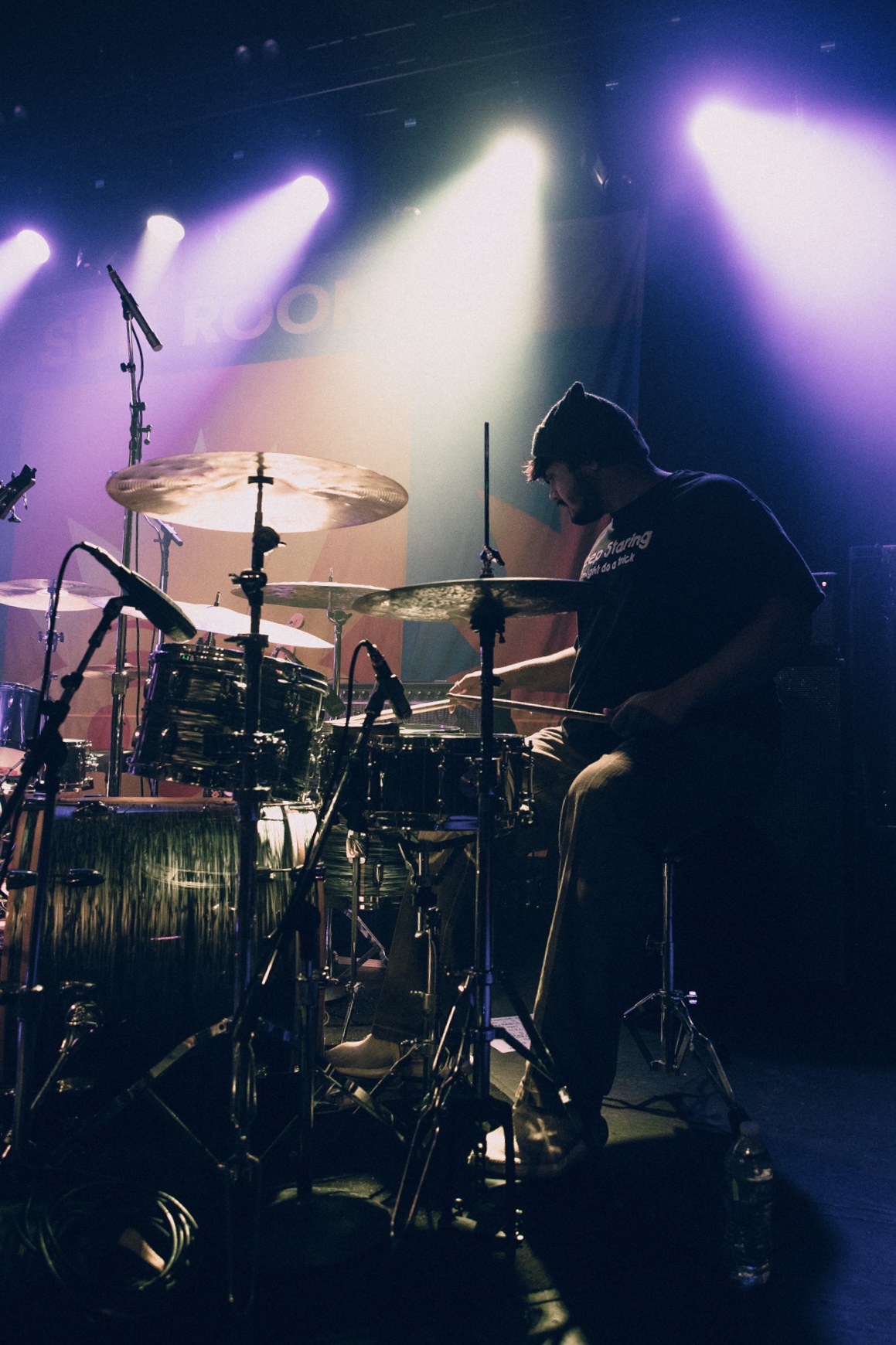 Silhouette of a drummer playing on stage with colorful lights shining behind, drums and cymbals visible.