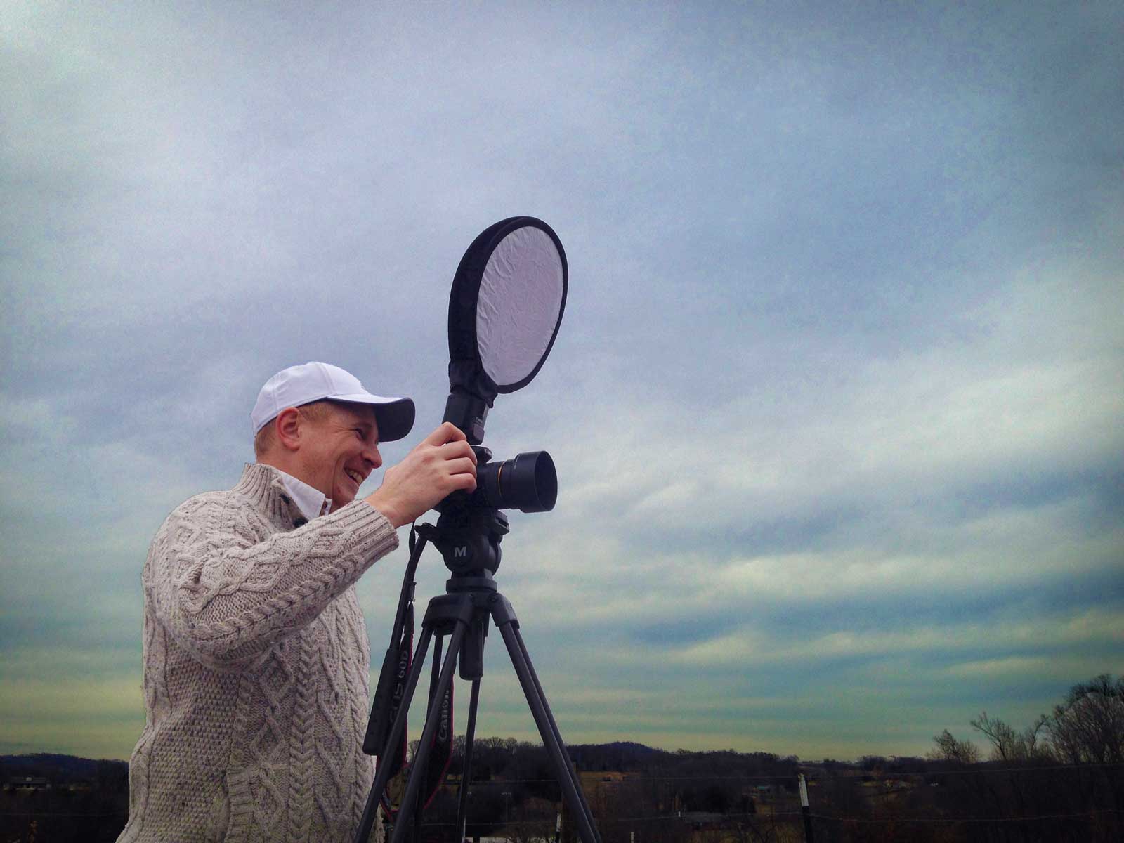 Person wearing a white cap and beige sweater, holding a camera with a large circular reflector, outdoors under cloudy sky