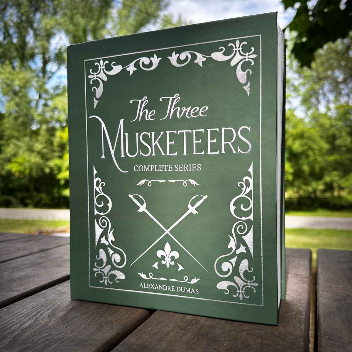 Book titled 'The Three Musketeers' by Alexandre Dumas, with decorative border and crossed swords on cover, outdoors on a wooden surface.