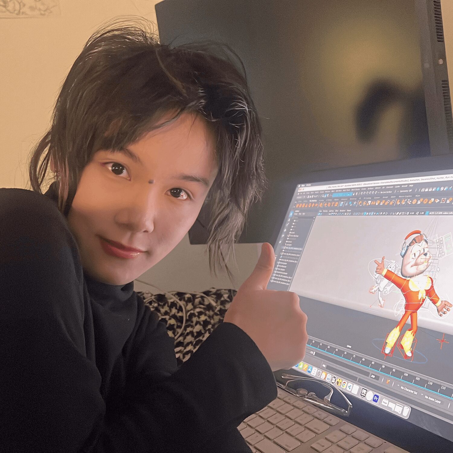 Person with short dark hair giving a thumbs-up, working on a computer with a cartoon character on the screen.