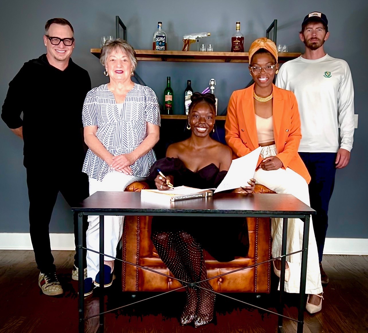 Five people, four standing and one sitting, in a room with a shelf of bottles behind them. The seated person is at a desk with a book and pen.