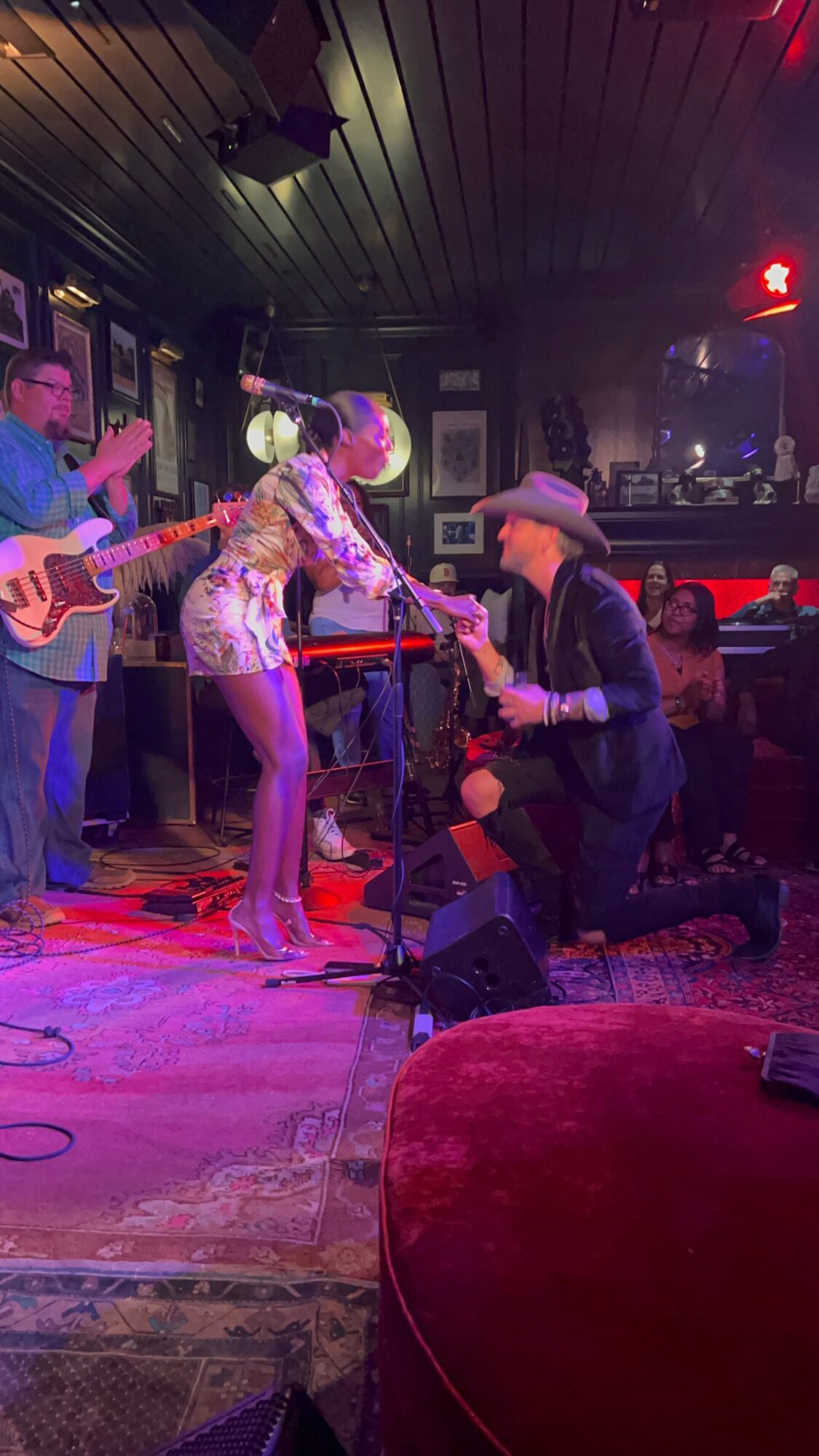 A woman in a floral shirt and a man in a cowboy hat are performing on stage with a guitarist in the background.
