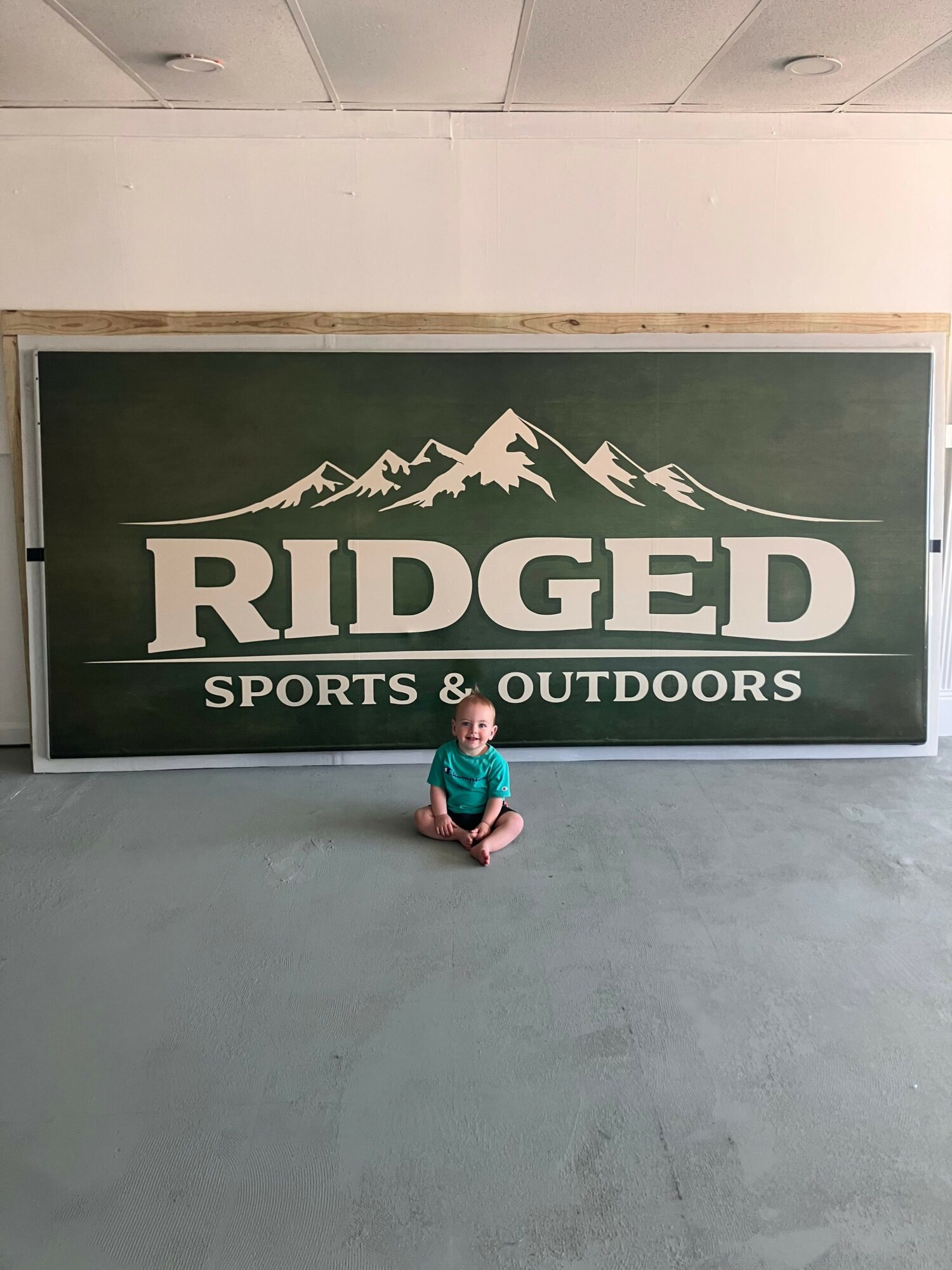 Child sitting on the ground in front of a large Ridge Sports & Outdoors sign with mountains, on a concrete floor.