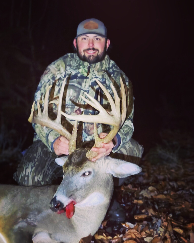 Man in camouflage clothing holding a large deer with antlers, outdoors at night, smiling, with a dark background.