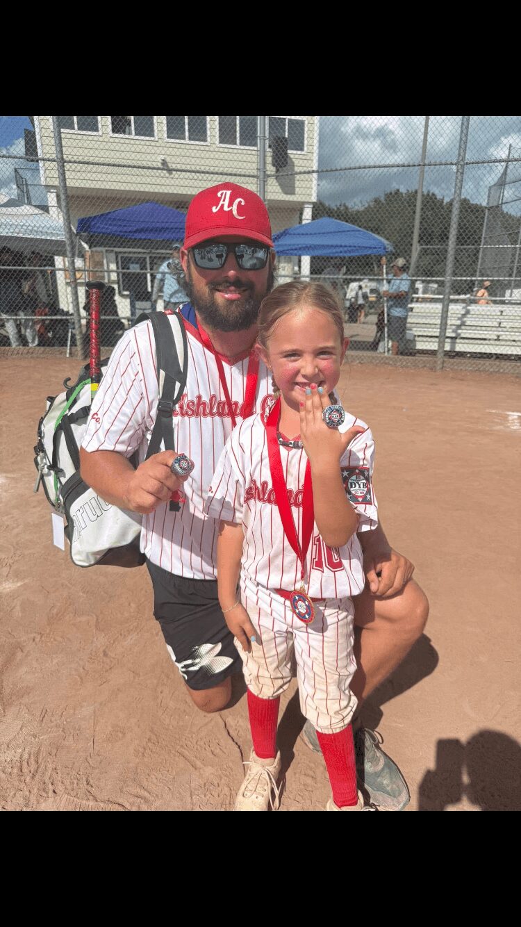 A man and a young girl in baseball uniforms on a baseball field, smiling, with a chain-link fence and tents in background.