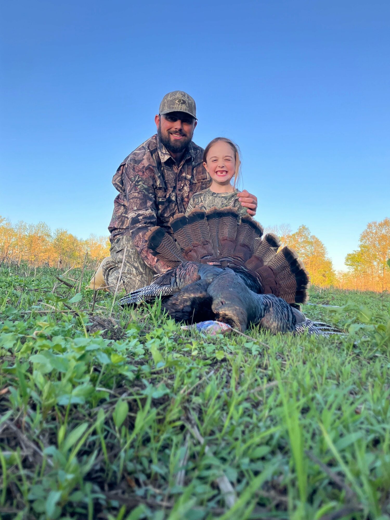 Two people in camouflage clothing kneel on grass with a turtle, trees, and a blue sky in the background.
