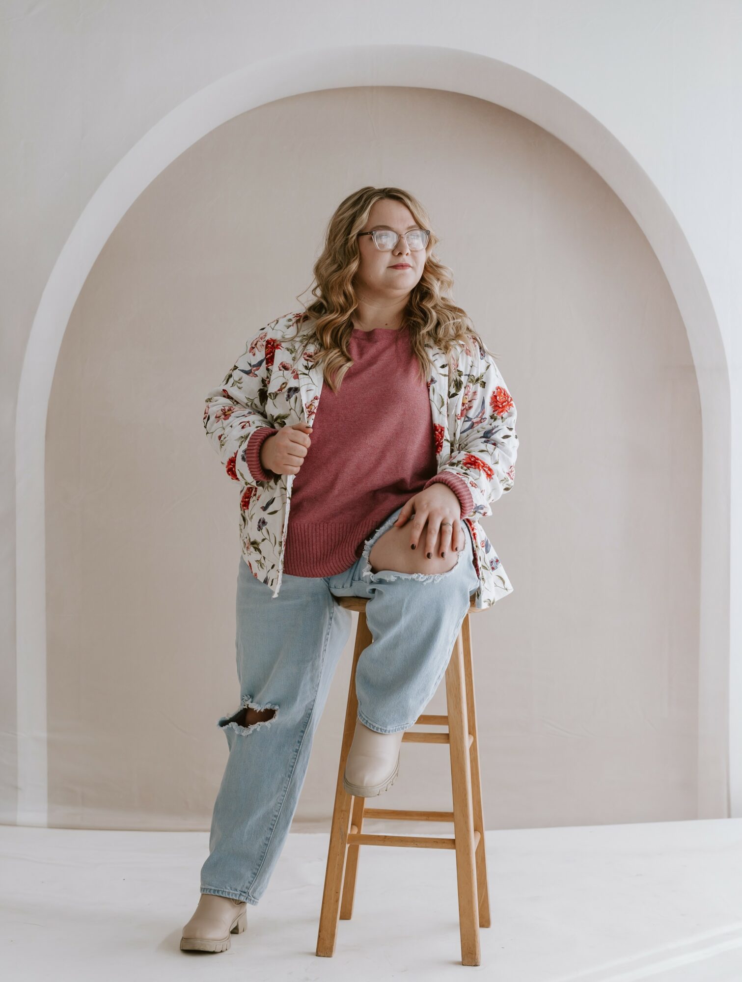 Woman with glasses and wavy hair, wearing a floral jacket, maroon shirt, and ripped jeans, sitting on a stool under an arch.
