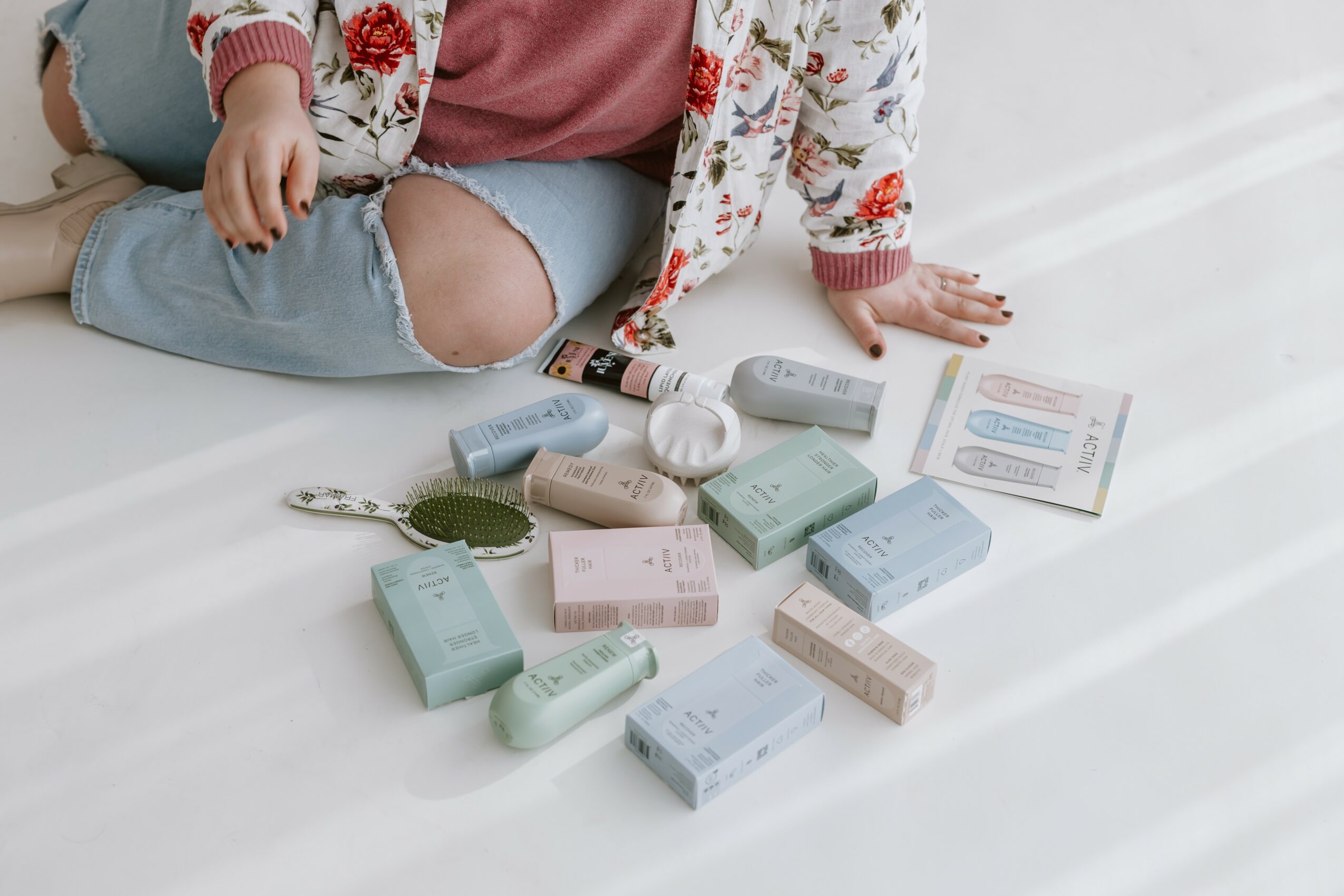 Child sitting on floor with various skincare and cosmetic products spread out, some open, on a white surface.