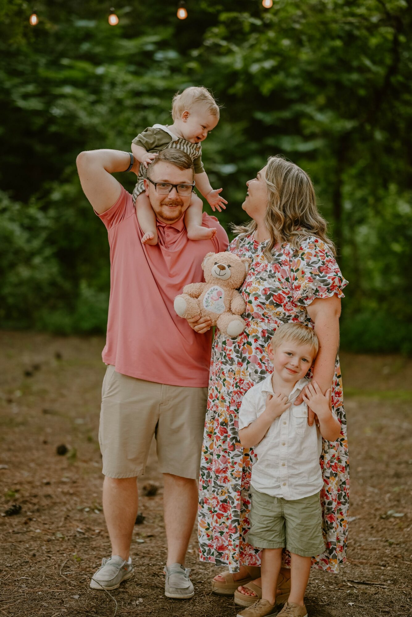 Family of four outdoors with trees in background, a man holding a child on his shoulders, a woman holding a teddy bear, and a young girl smiling.