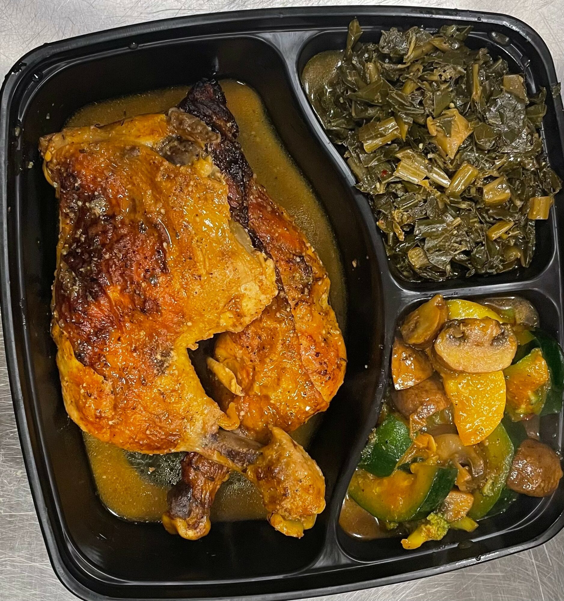 Black tray with baked chicken, cooked greens, and vegetable stew, divided into three sections.