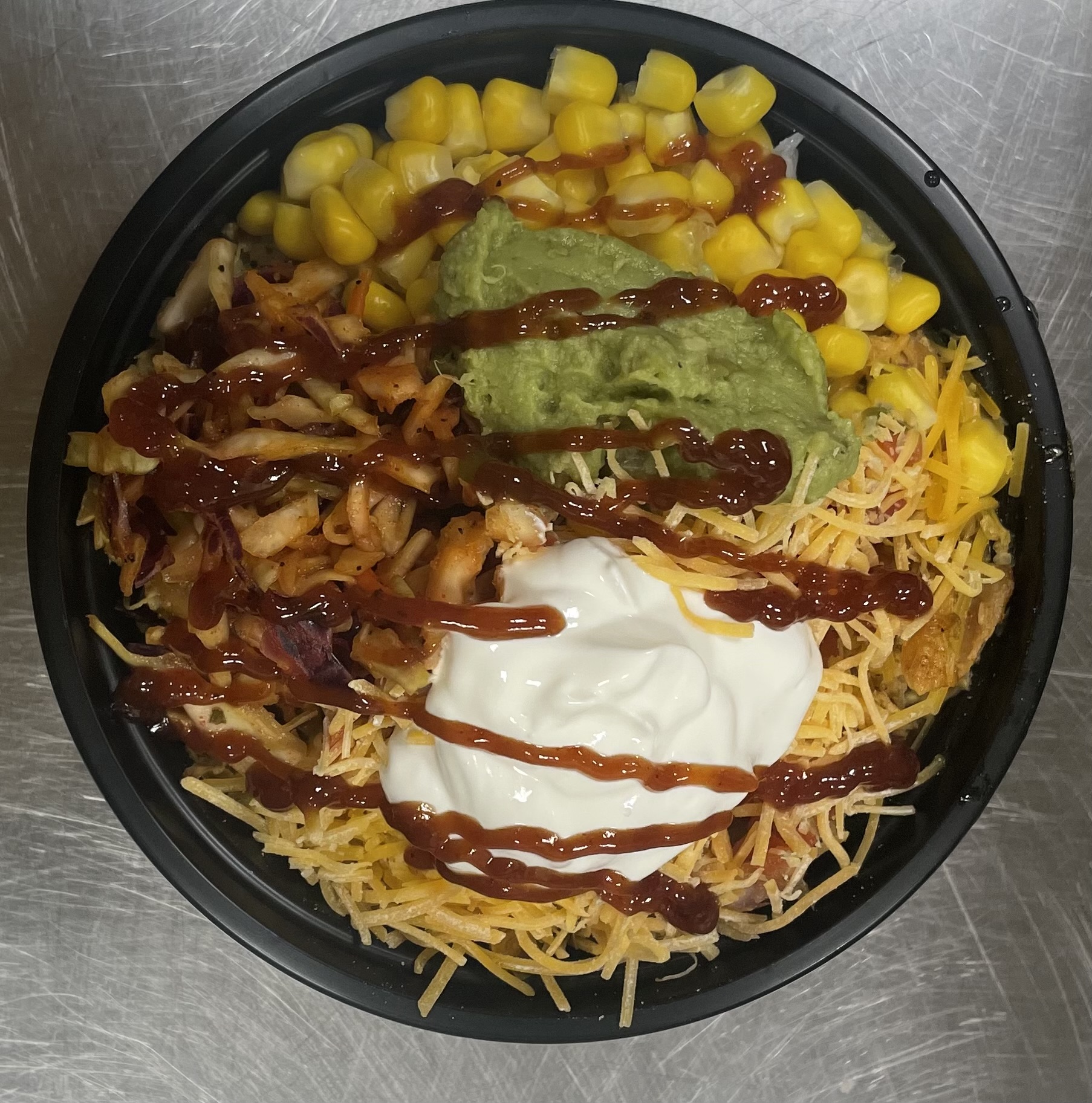 Bowl of shredded cheese, corn, guacamole, sour cream, and ketchup on a metal surface.