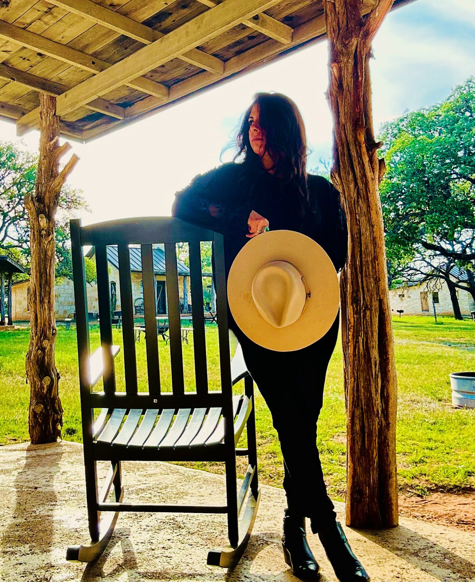 Person holding a wide-brimmed hat standing outdoors near a wooden chair, with trees and a grassy area in the background.