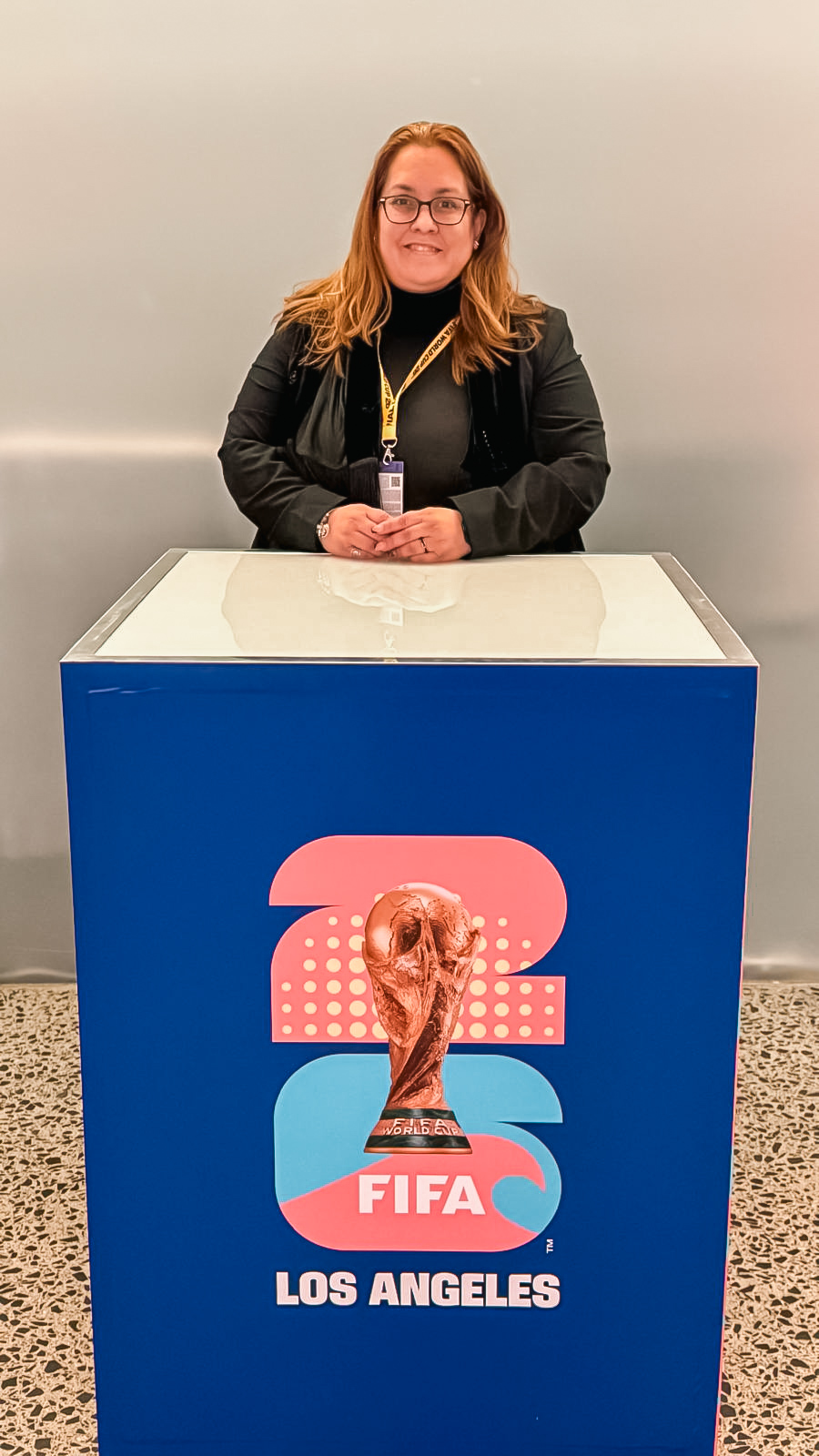Woman with glasses and long hair standing behind a FIFA Los Angeles display, smiling, against a plain wall.