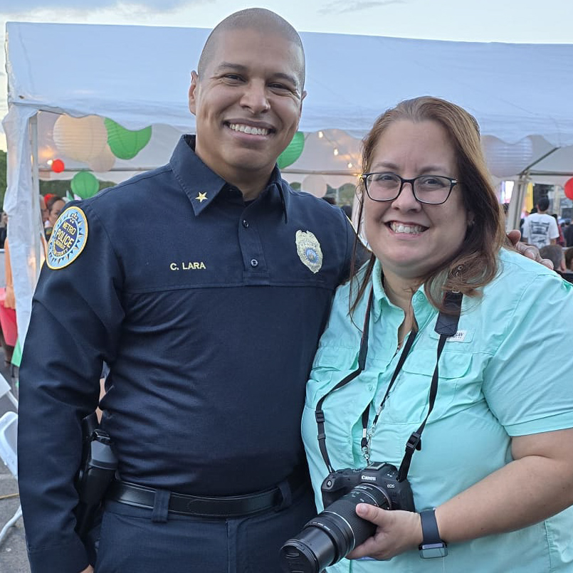 Two smiling women, one in police uniform and the other with a camera, standing outdoors under a tent.