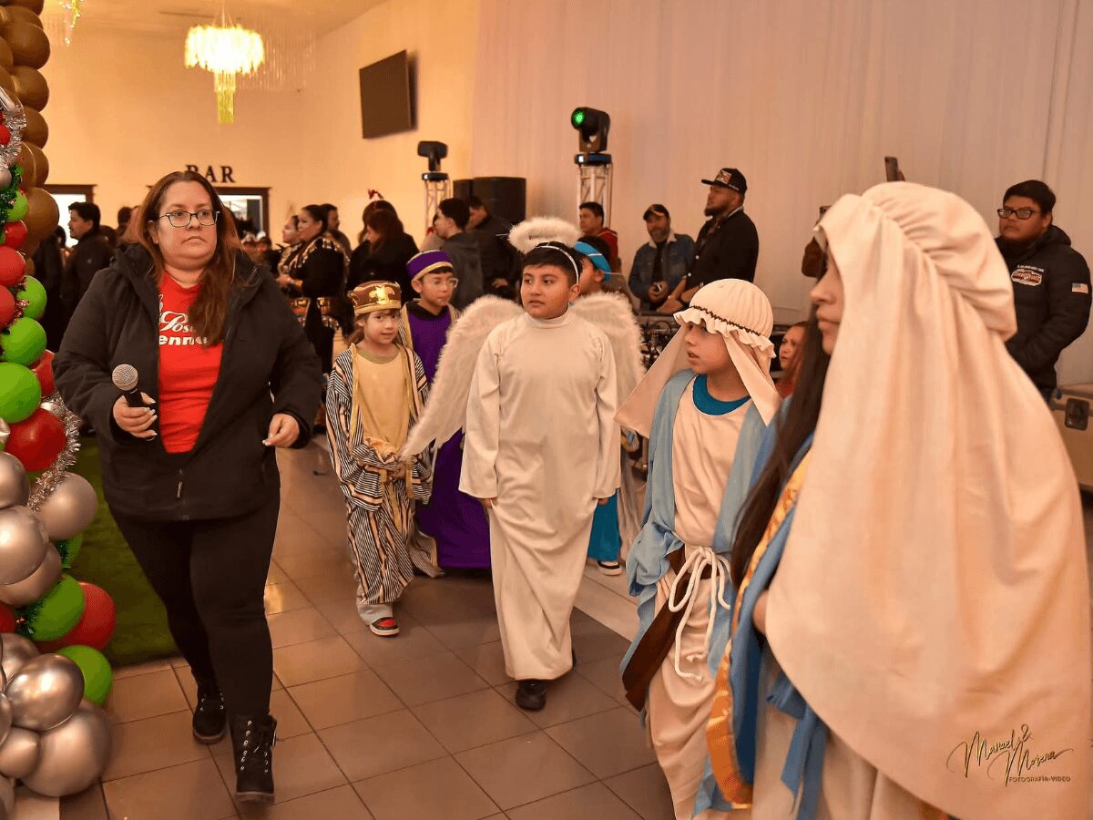 Children dressed in nativity costumes walk in a line at a festive event, with adults and decorations in the background.