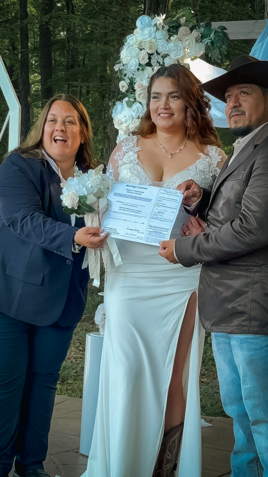 Three people at a wedding ceremony outdoors, holding a document, smiling, with floral decorations in the background.