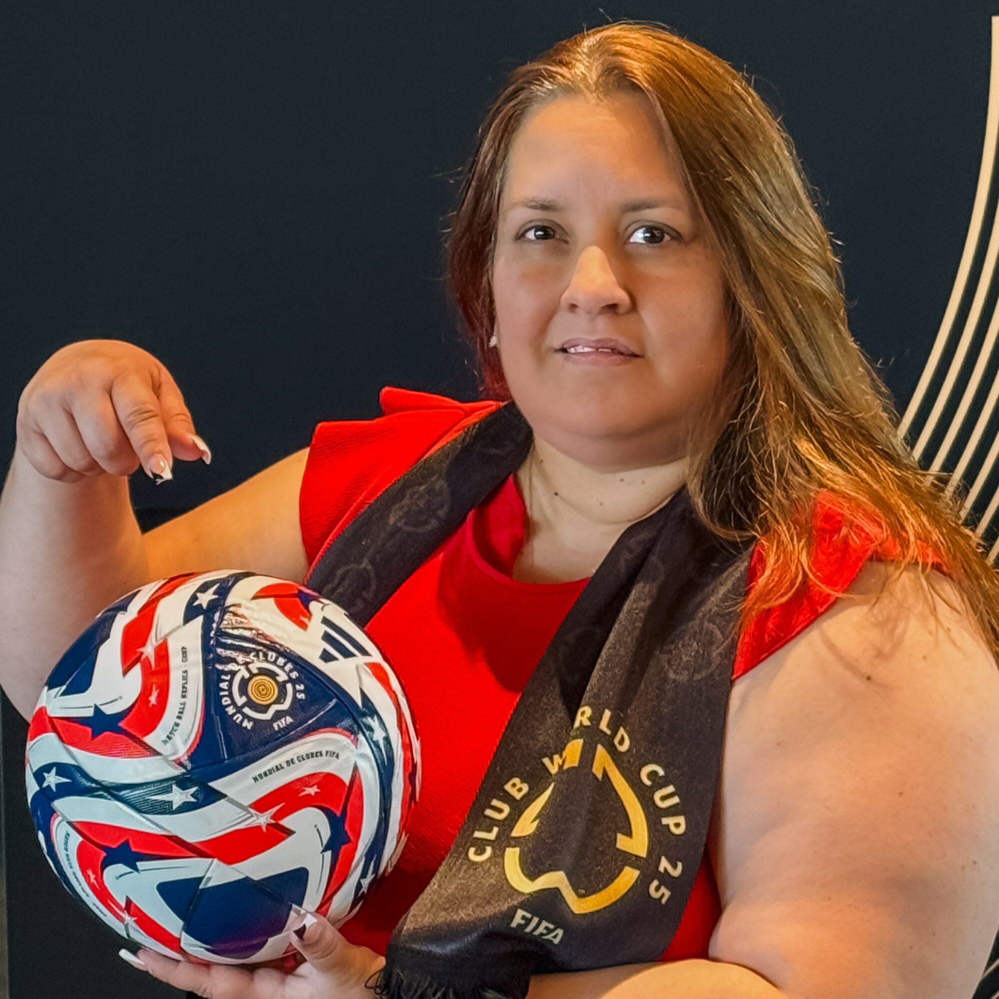 Woman holding a colorful soccer ball, wearing a red shirt and a black scarf with yellow logo, against a dark background.