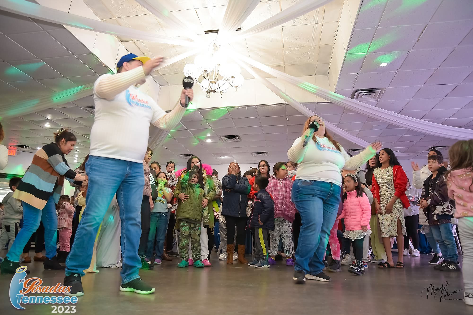 People gathered in a large indoor space with two performers in white shirts and blue pants, colorful lights, and a crowd of children and adults.