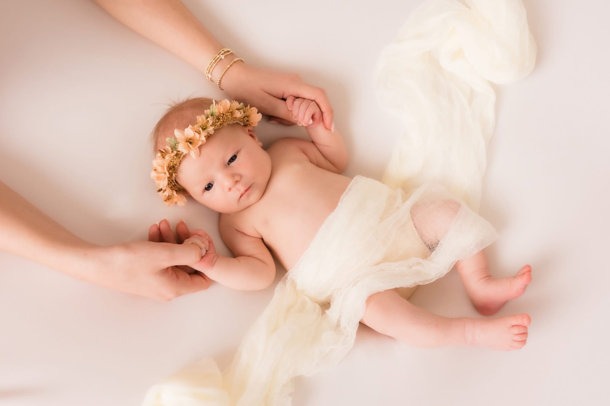 Baby lying on a soft surface, wearing a flower crown and a light cloth, holding hands with an adult's hands.