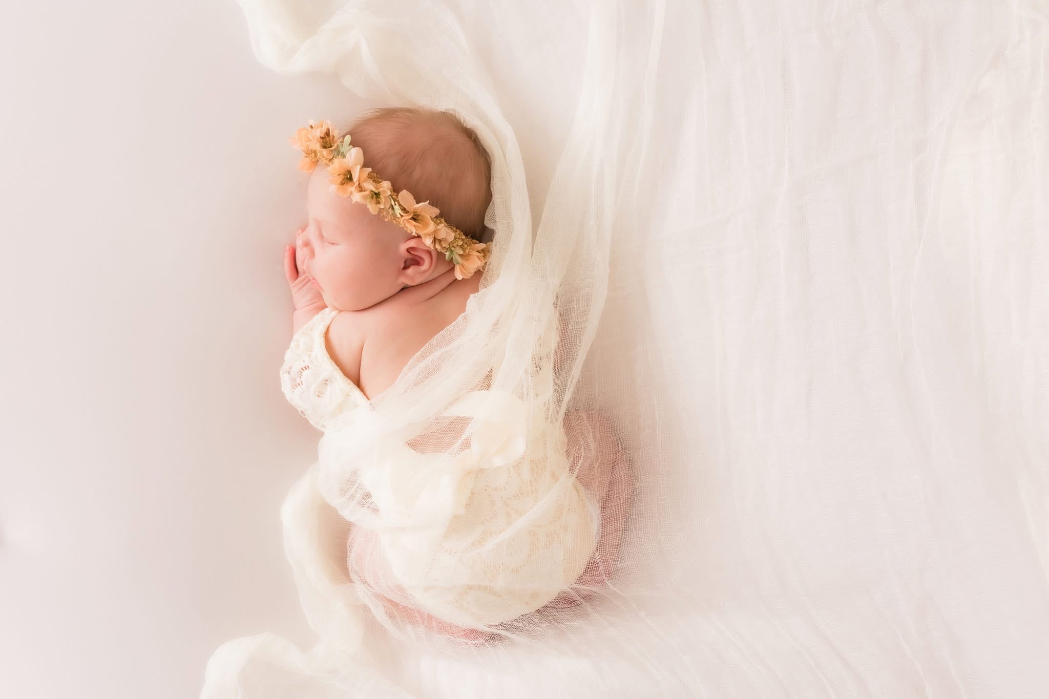 A sleeping newborn baby with a floral headband, lying on a soft white blanket, wrapped in a light fabric.