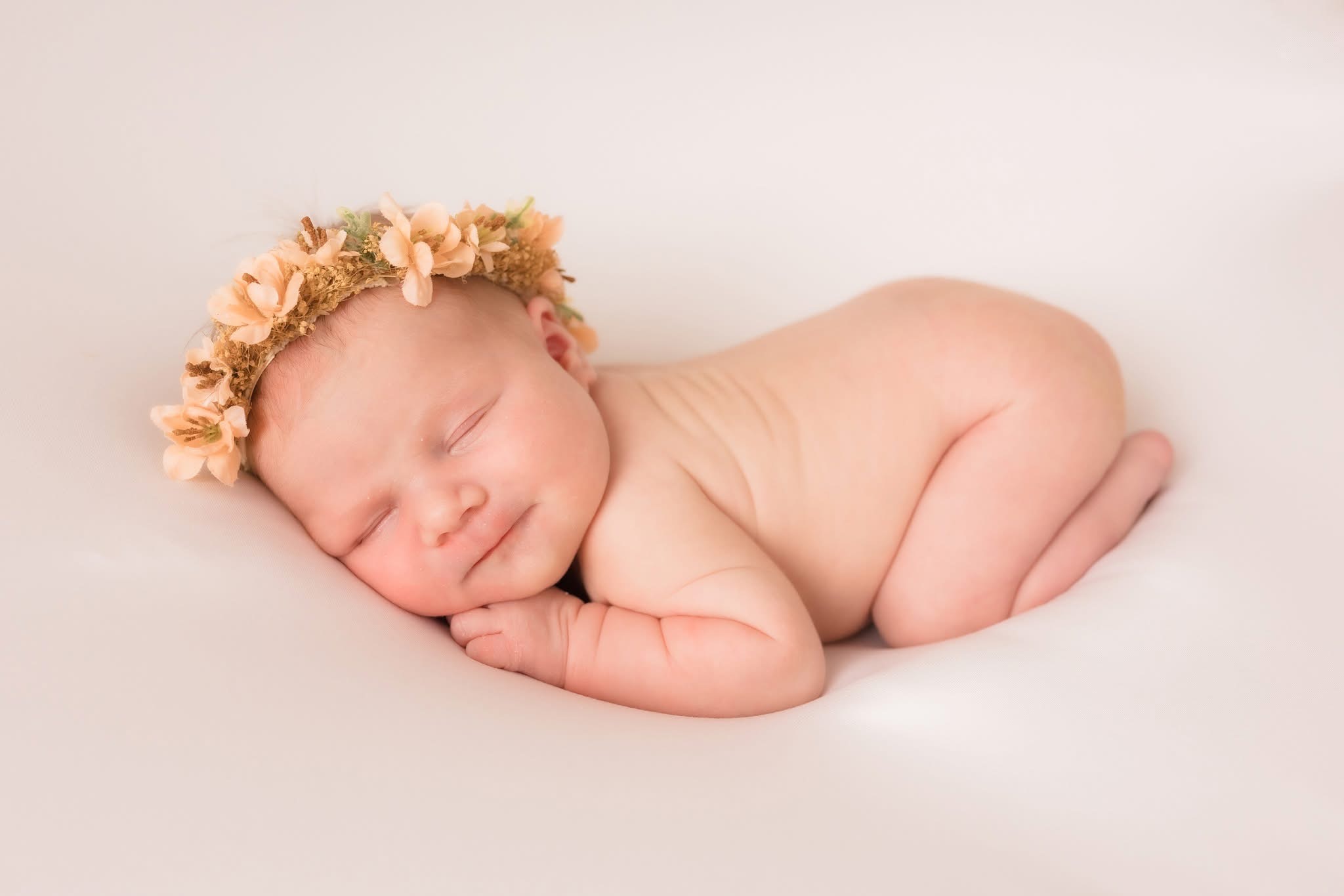 Sleeping baby with a flower crown, lying on side with hands under face, on a white surface.