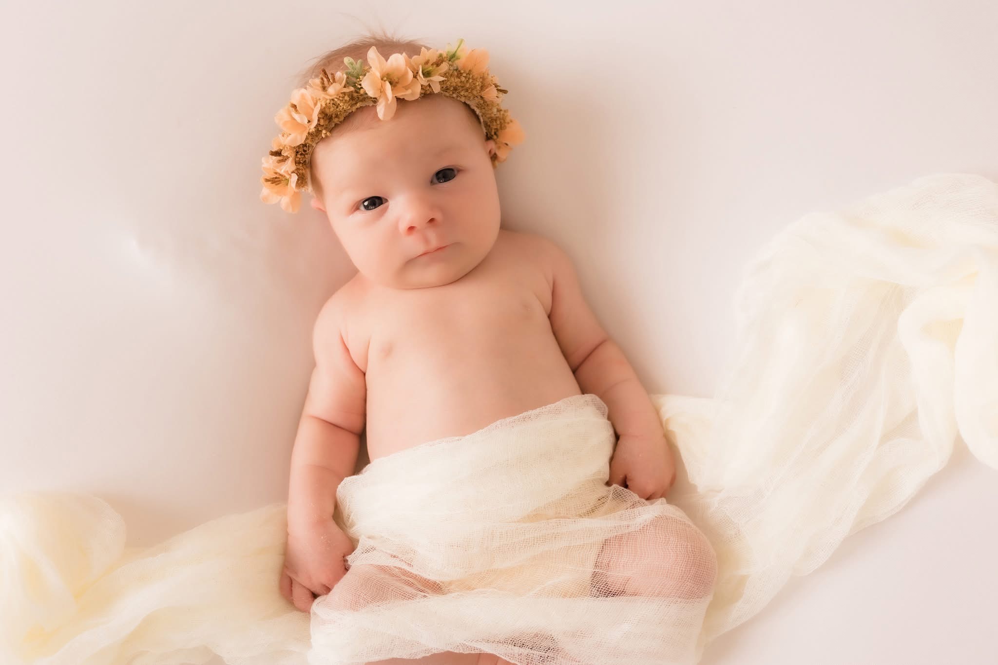 Baby with a flower crown sitting on a soft surface, wrapped in a light cloth, looking at the camera.
