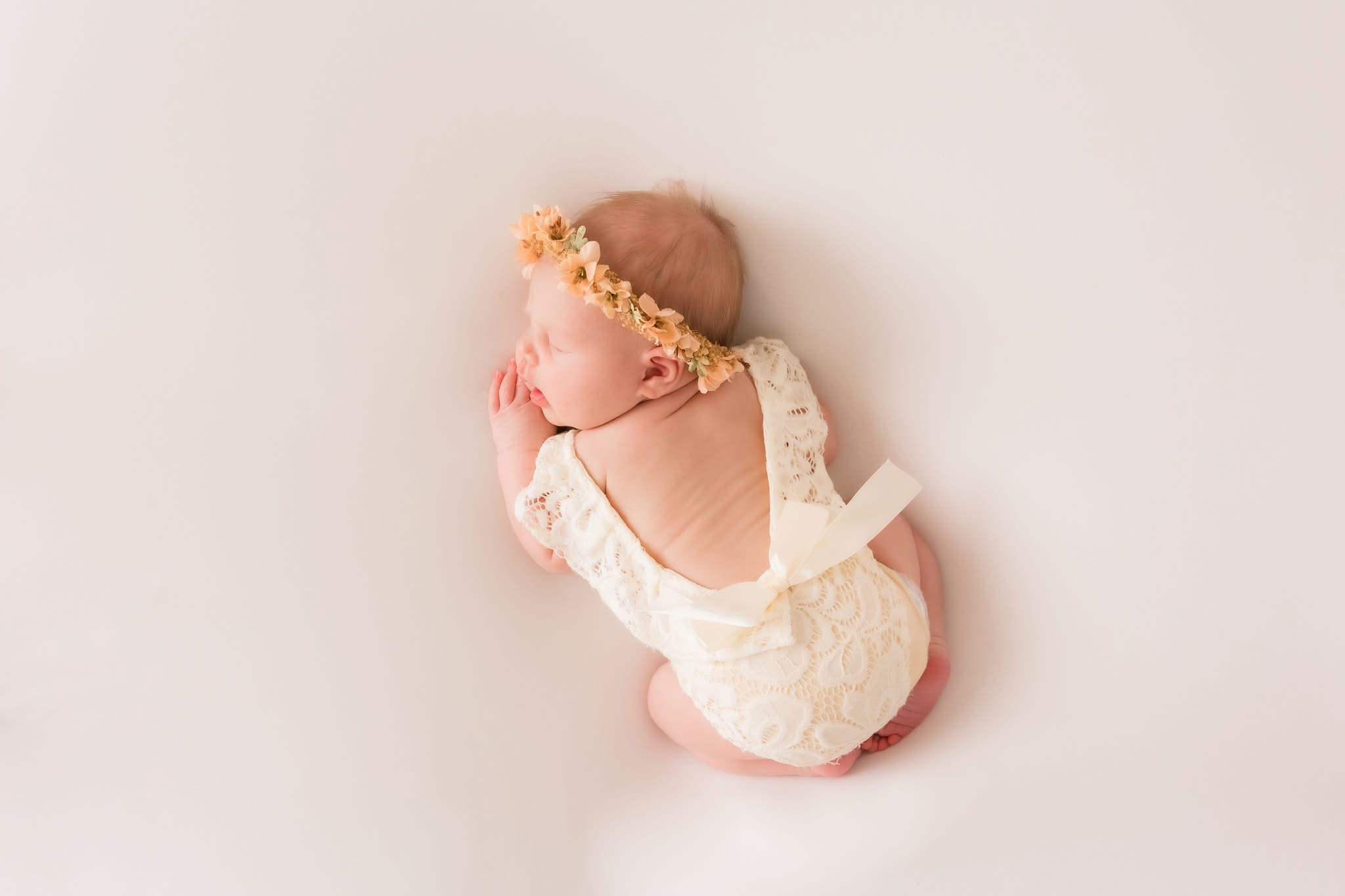 Newborn baby sleeping on side, wearing a floral headband and lace outfit, on a plain light background.
