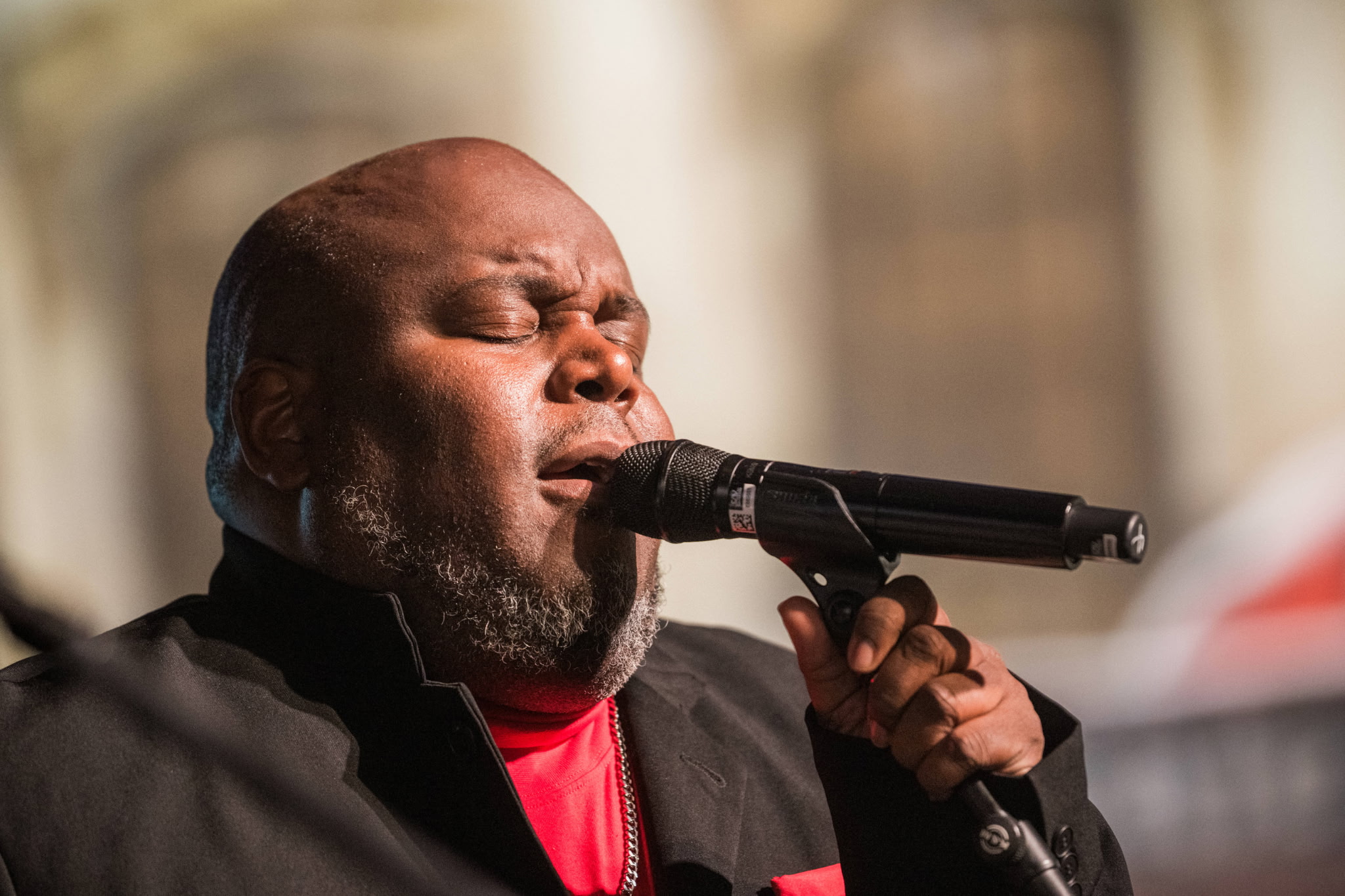 A man with closed eyes singing into a microphone outdoors, wearing a black jacket and red shirt.