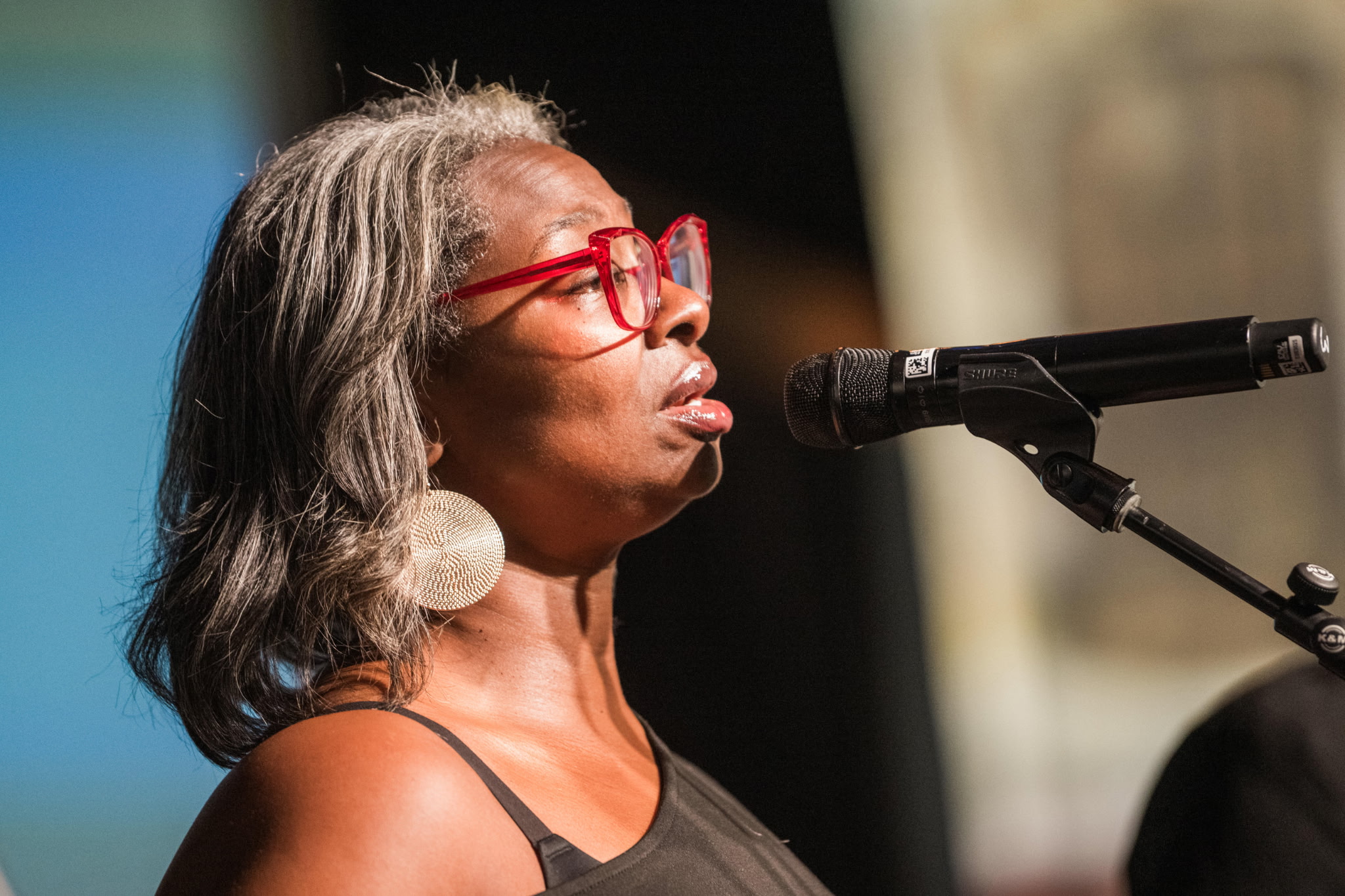 Woman with gray hair, red glasses, and large earrings speaking into a microphone, wearing a black top.