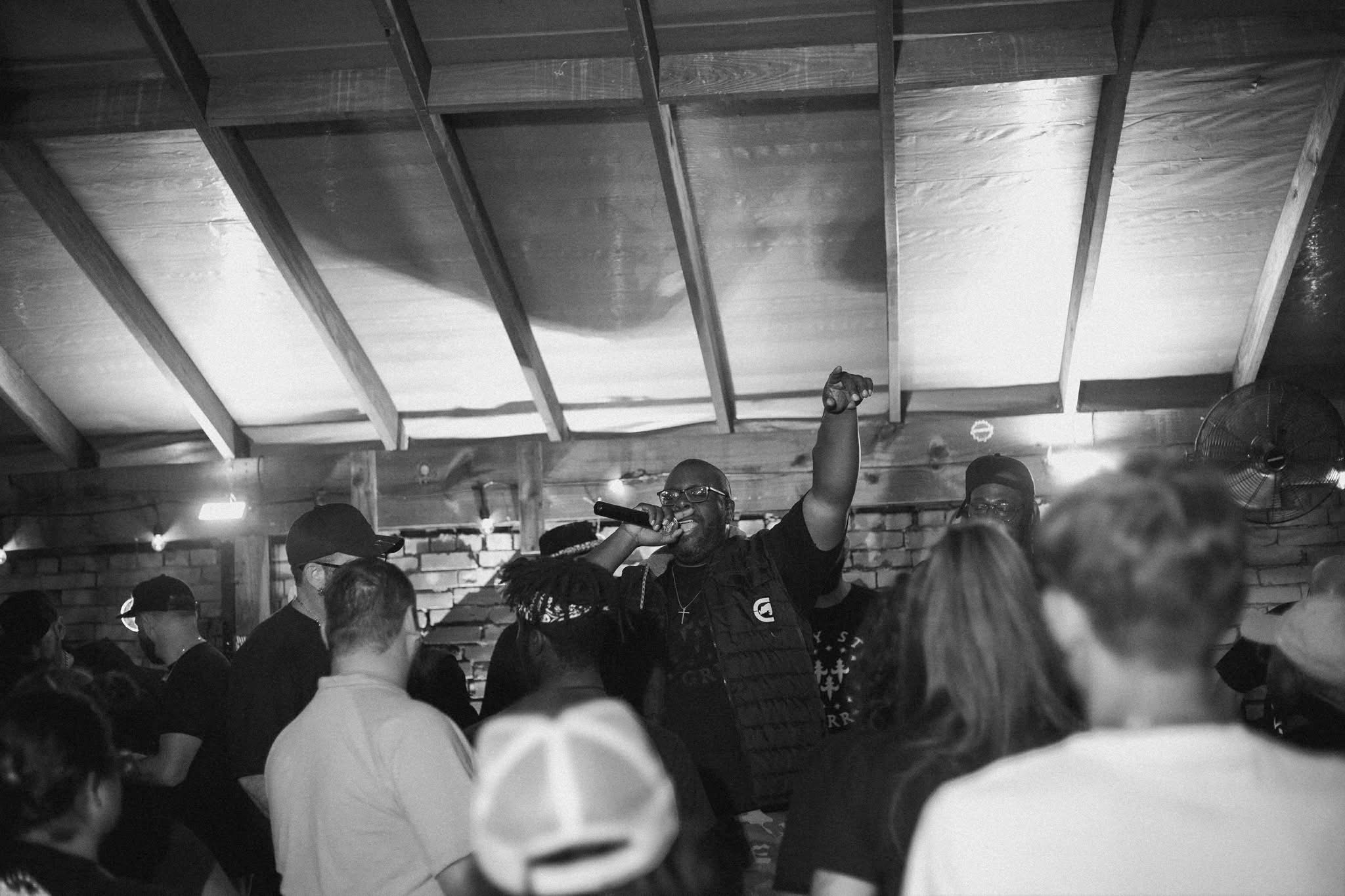People gathered in a dimly lit indoor space with a person raising a fist in the center, ceiling with wooden beams visible.