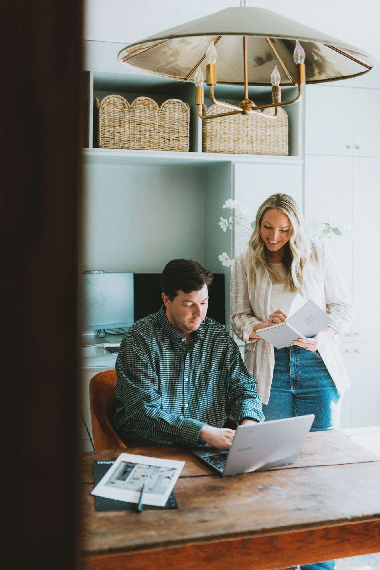 Two people working together in a bright office, one seated at a desk with a laptop, the other standing with papers.