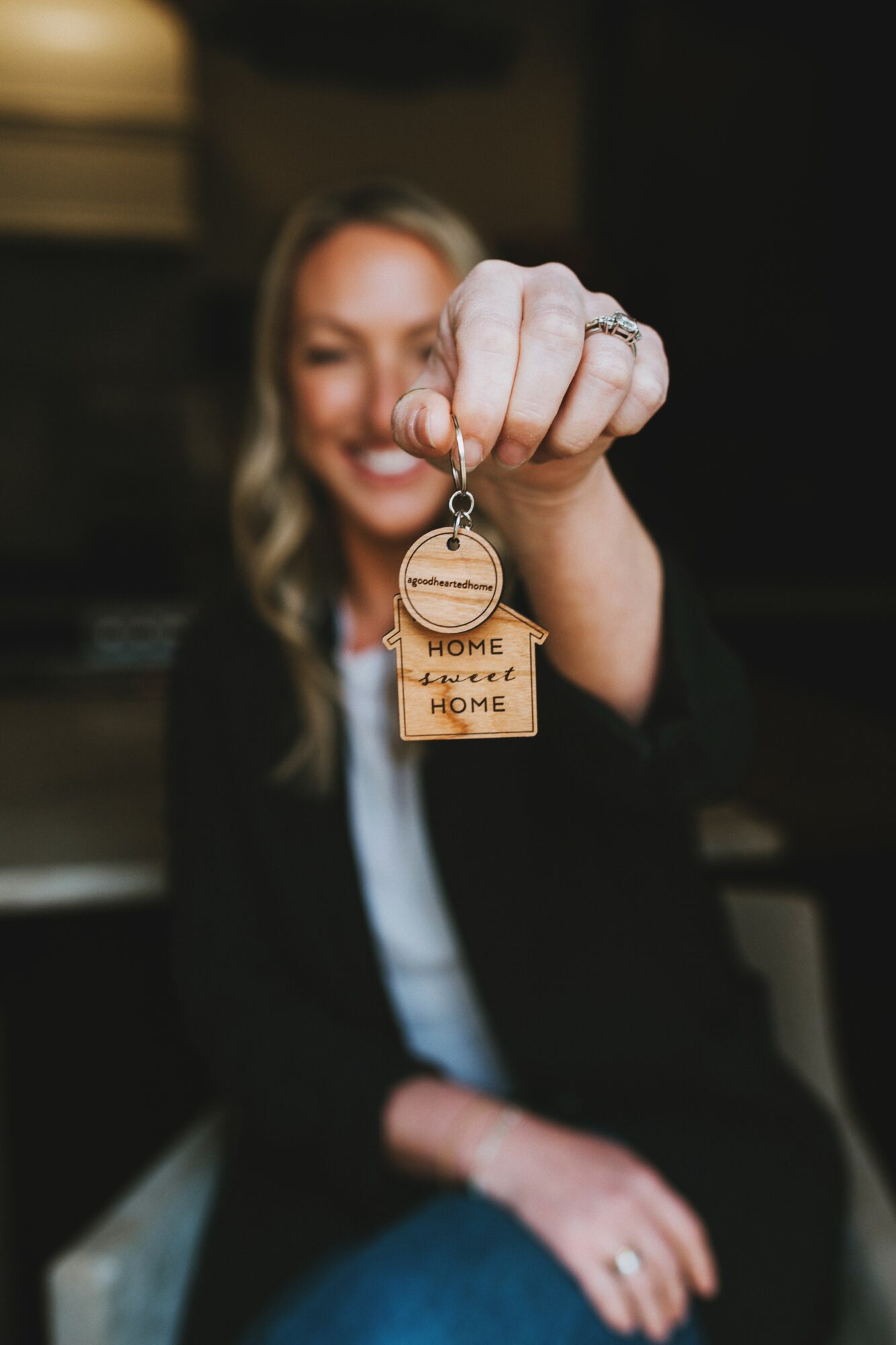 Woman holding a keychain with a house and 'HOME' text, smiling, blurred background, focus on hand and keychain.
