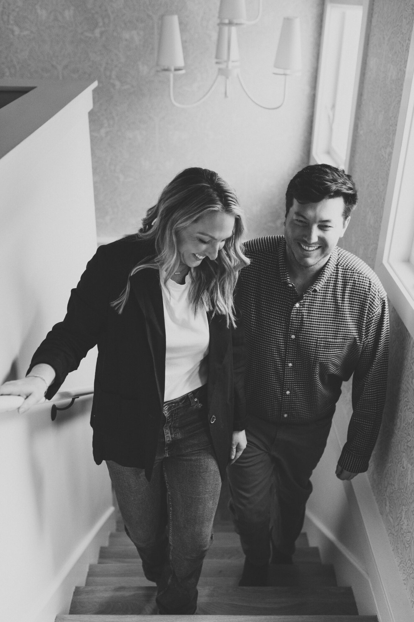 Two people walking up stairs indoors, smiling and talking, with a wall and window nearby.