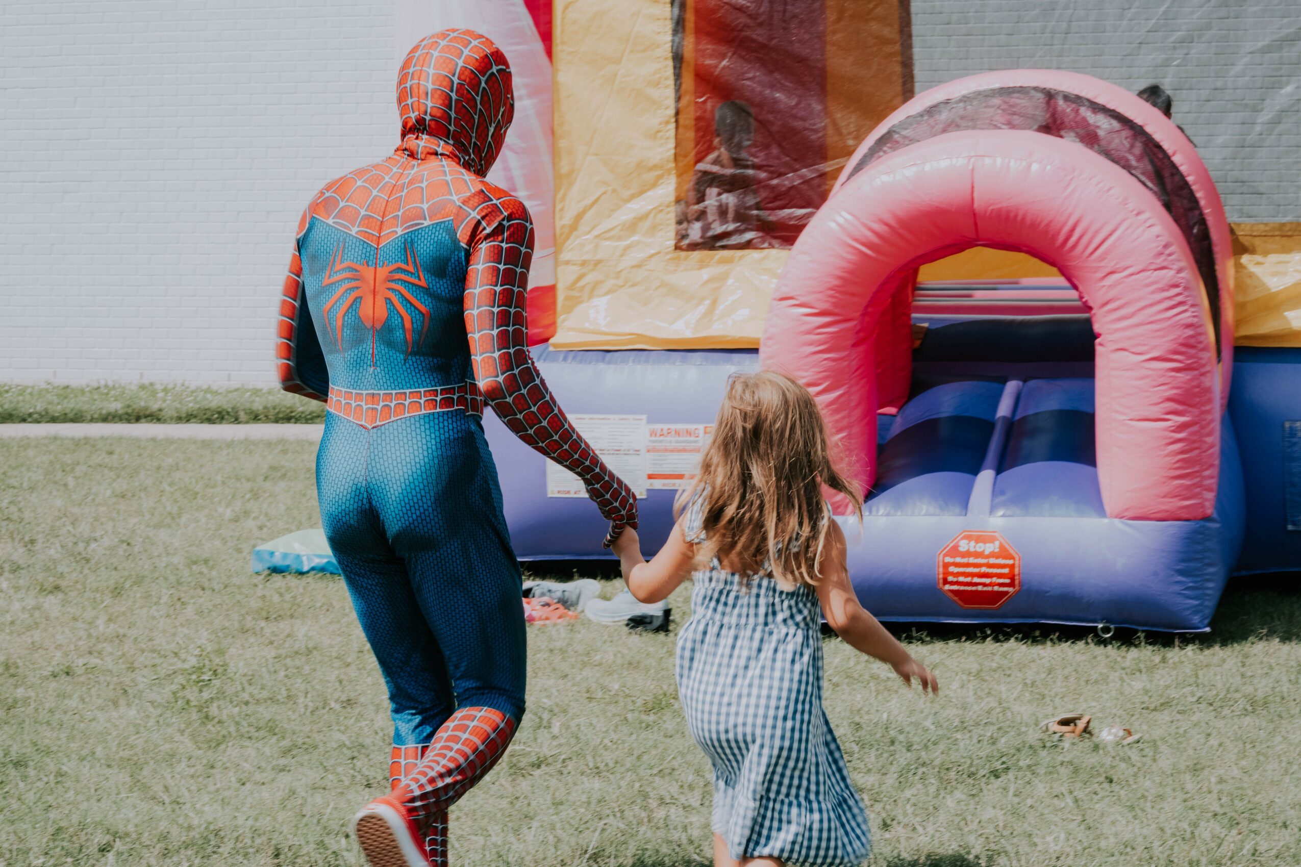 Person dressed as Spider-Man holding hands with a young girl near an inflatable slide at an outdoor event.
